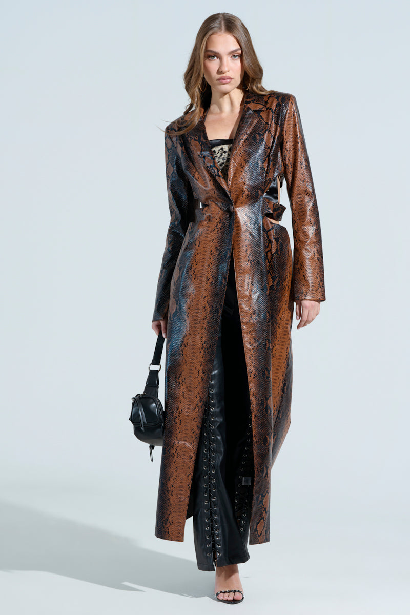 A woman stands confidently against a light background, wearing the MORE LIFE CUTOUT FAUX LEATHER TRENCH, black pants with lace-up details, and black heels. She carries a small black bag and lets her hair fall loose.