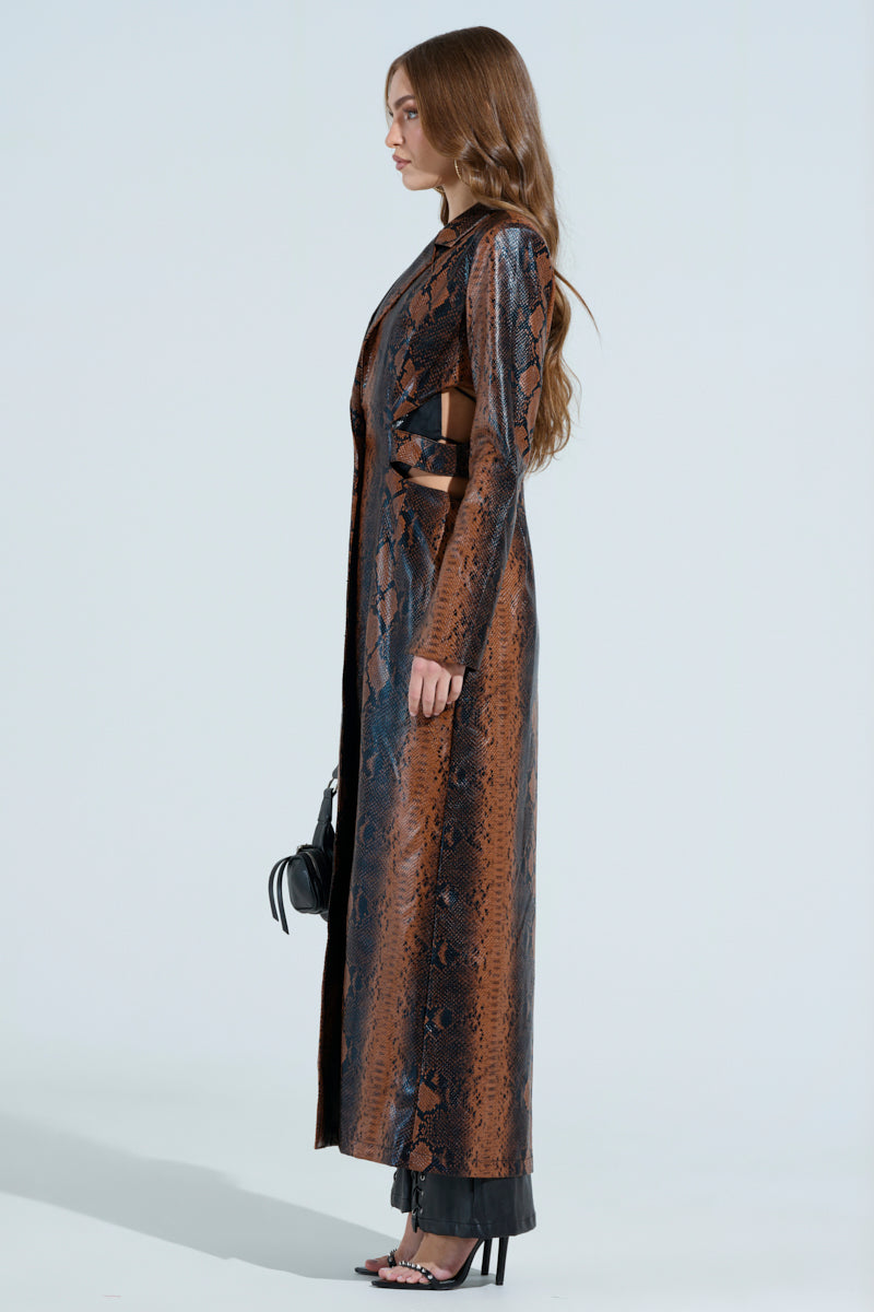 Against a neutral background, a woman with long wavy hair wears the chic MORE LIFE CUTOUT FAUX LEATHER TRENCH, showcasing its snakeskin pattern. She pairs it with a small black handbag and matching shoes.