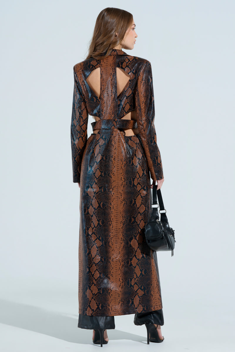 A person with long hair wears the MORE LIFE CUTOUT FAUX LEATHER TRENCH in a brown snakeskin pattern with cut-out faux leather details on the back. They hold a black handbag, wear black shoes, and stand against a plain light gray background.