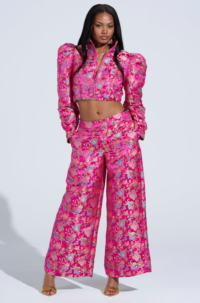 In a plain white setting, a woman exudes confidence wearing the vibrant JULES BROCADE TROUSER with its floral pattern and wide-leg design, complemented by puffed sleeves, a cropped jacket, and gold-heeled sandals.