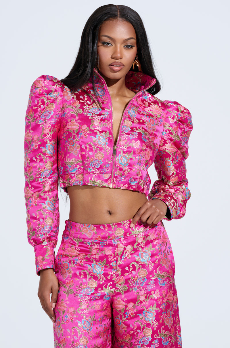 A woman wears the vibrant pink and gold JULES BROCADE BOMBER with puff sleeves and matching high-waisted pants. Her long black hair cascades against a plain white background, adding elegance to the look enhanced by a subtle metallic zipper closure.