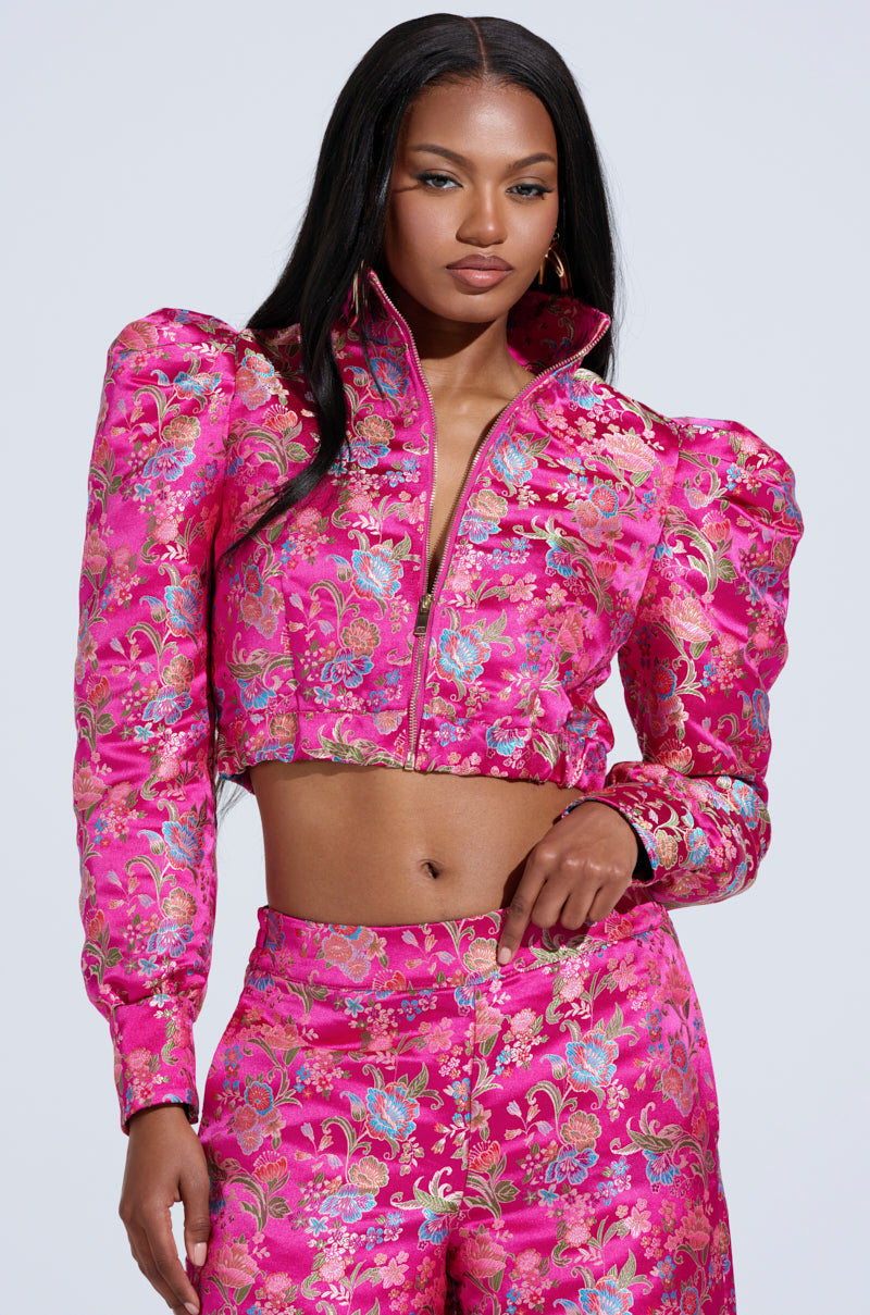 She wears the vibrant JULES Brocade Bomber with a floral brocade pattern and puffed shoulders, complemented by matching high-waisted pants. Her sleek, straight black hair enhances her confident expression, while the bomber's metallic zipper closure stands out against a plain light background.