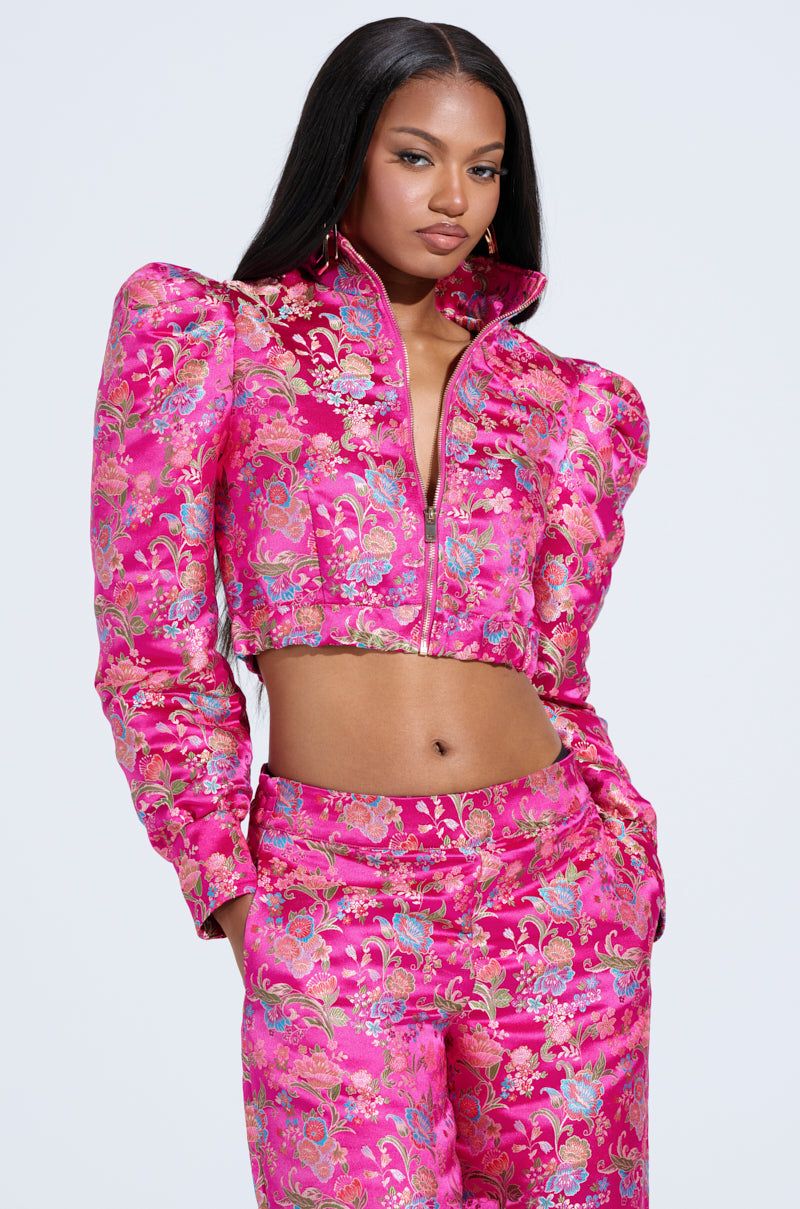 In a vibrant pink JULES Brocade Bomber adorned with gold and silver floral patterns, featuring puffed shoulders and a metallic zipper, one dazzles in style while pairing it effortlessly with matching high-waisted pants against a plain backdrop.