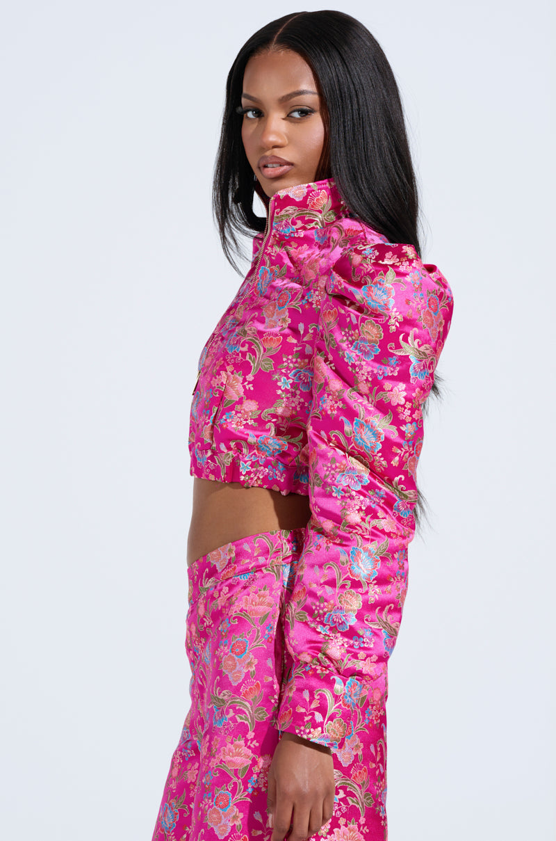 A woman wears the JULES BROCADE BOMBER, a vibrant pink, floral brocade-patterned long-sleeve crop top with a metallic zipper closure and matching high-waisted skirt. She has long, straight hair and gazes confidently to the side against a white background.