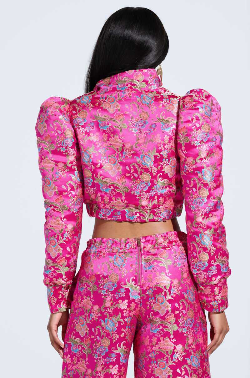 A person with long dark hair wears a vibrant pink JULES Brocade Bomber with intricate floral embroidery and puffed shoulders, paired with matching high-waisted pants. The metallic zipper closure adds a modern touch to the look against a light blue background.