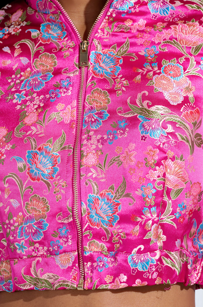 This close-up highlights the JULES BROCADE BOMBER, a luxurious vibrant pink satin jacket with an intricate floral brocade pattern in blue, gold, and pink. It features elaborate embroidery and a striking metallic zipper closure.