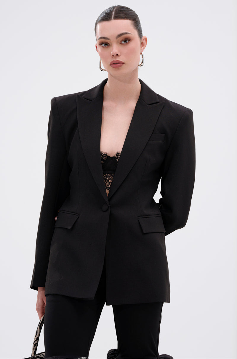 A woman with slicked-back hair wears the PERFECT FIT BLAZER IN BLACK, featuring a notched lapel collar over a lace top and accessorized with large hoop earrings. She poses confidently against a light background, holding a bag in her left hand.