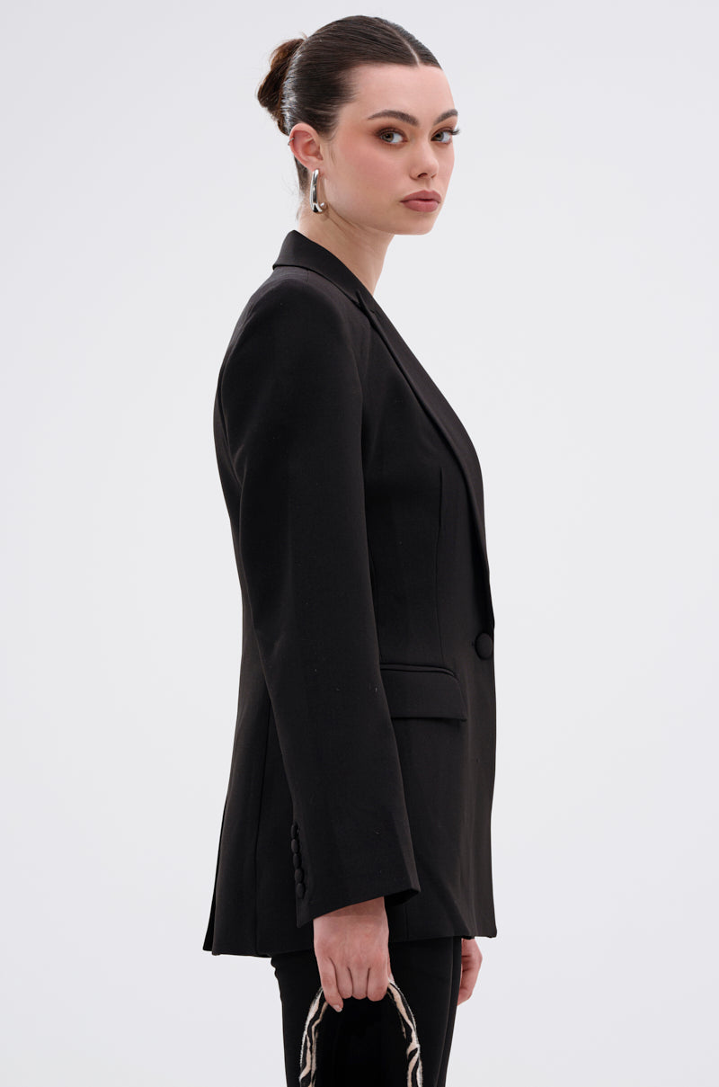 A woman with her hair in a bun, wearing the PERFECT FIT BLAZER IN BLACK with a notched lapel collar and holding a small handbag, stands sideways and looks over her shoulder against a plain light background.