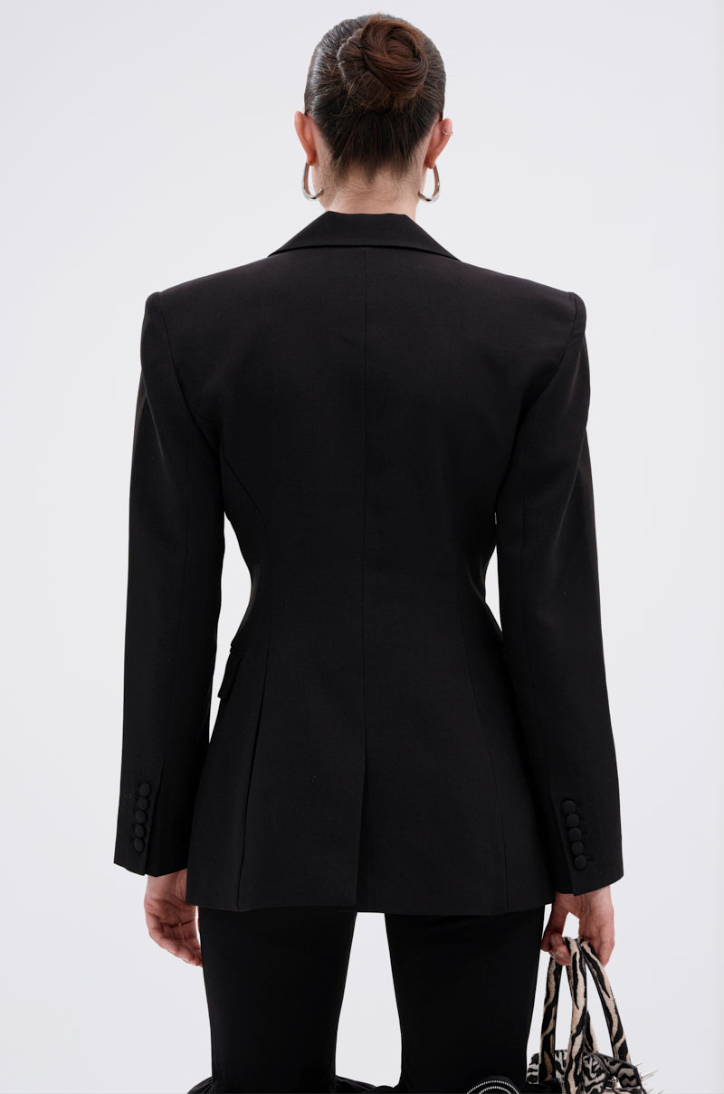 A woman with her hair in a neat bun wears the PERFECT FIT BLAZER IN BLACK paired with black pants. Seen from the back, she holds a patterned handbag in her left hand against a plain light background.