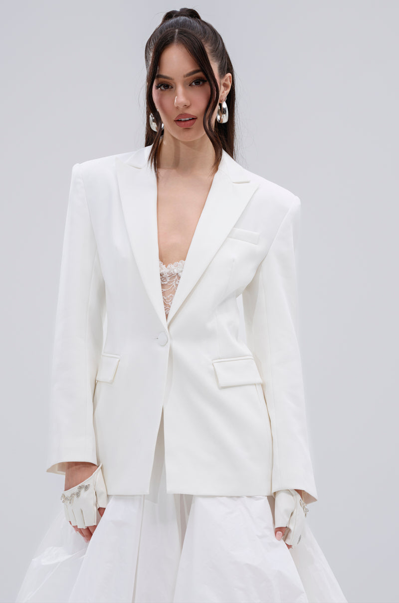 A woman with long dark hair styled in a half-up look wears the PERFECT FIT BLAZER over a lace top, white gloves, and a layered white skirt, posing against a plain light gray background.
