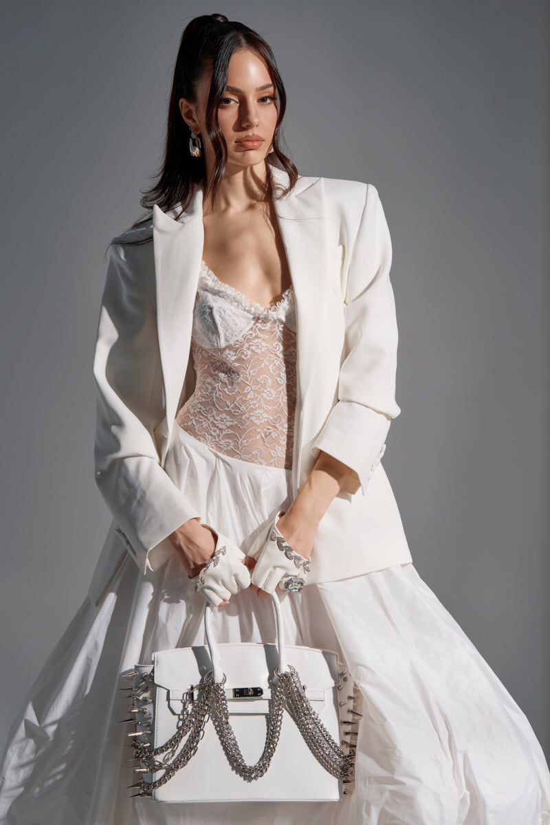 A woman in a white lace top, PERFECT FIT BLAZER, and flowing white skirt holds a white handbag with silver chains. She accessorizes with white fingerless gloves and silver jewelry, standing against a gray background.