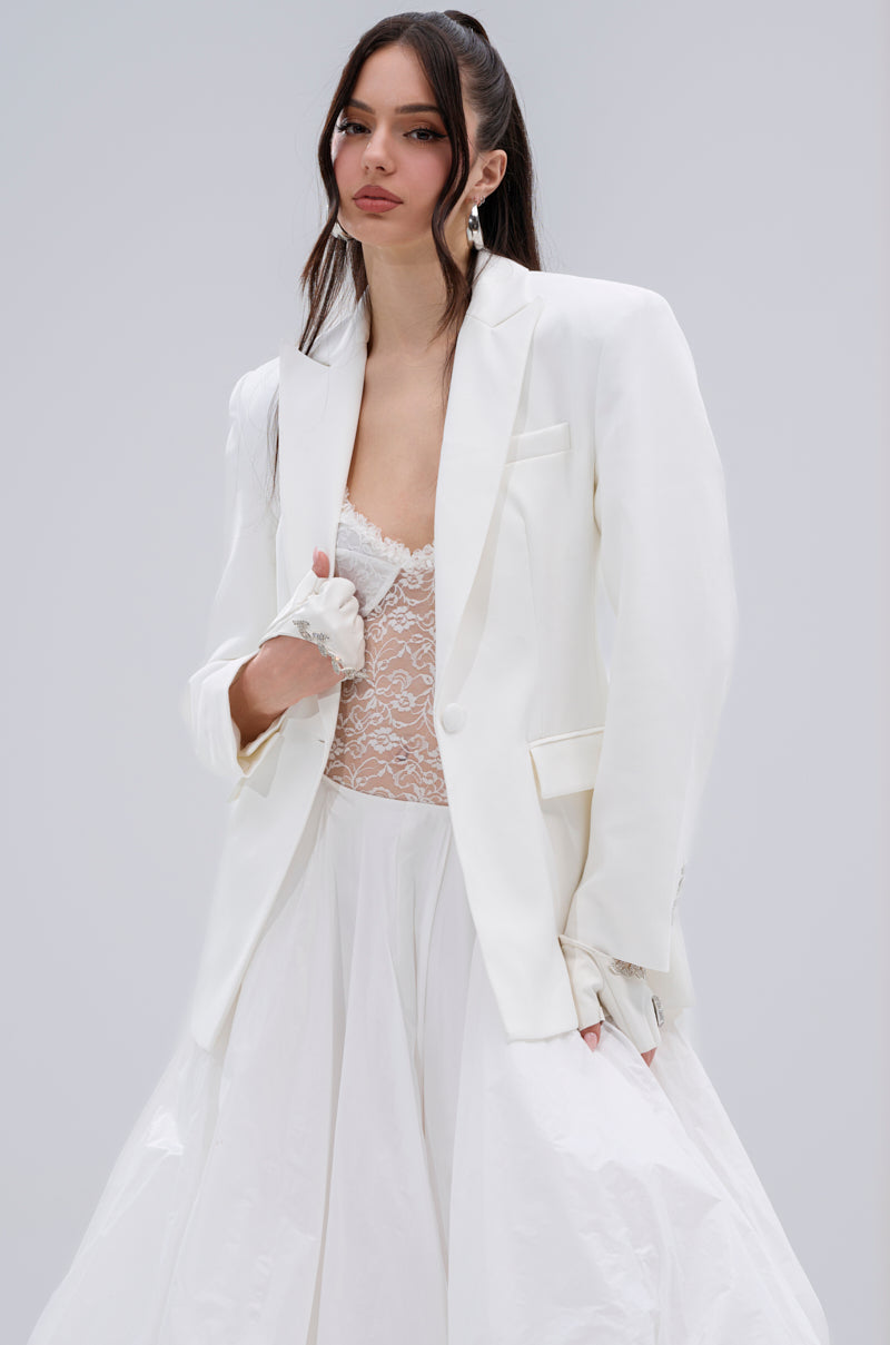 A woman with long, wavy dark hair and a confident look poses against a plain light grey background, wearing the PERFECT FIT BLAZER over a sheer lace top and a flowing white skirt.