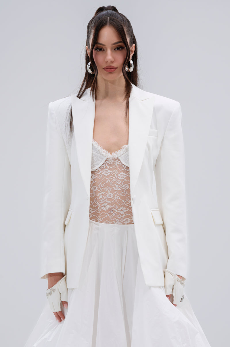 A woman with long dark hair models the PERFECT FIT BLAZER over a sheer lace top and flowing white skirt, set against a simple light background. She wears statement earrings and displays a confident look.