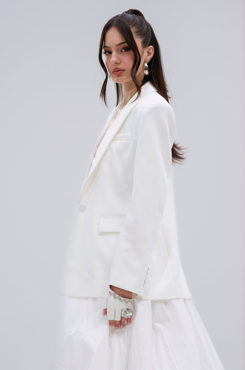 A woman with long brown hair in a half-up style models the PERFECT FIT BLAZER and a white skirt, styled with statement earrings and chunky rings, posing against a plain light gray background.