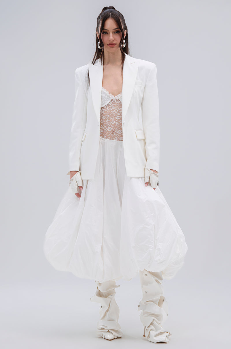 A woman poses against a plain backdrop in the PERFECT FIT BLAZER layered over a lace top, paired with a voluminous white skirt, white gloves, and tall sculptural boots. Her loose waves frame her face for an elegant look.