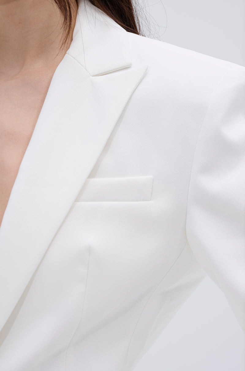 Close-up of a person wearing the PERFECT FIT BLAZER, made from sleek suiting material with a notched lapel and pocket details. Only the neck, shoulder, and chest are visible against a plain light background.