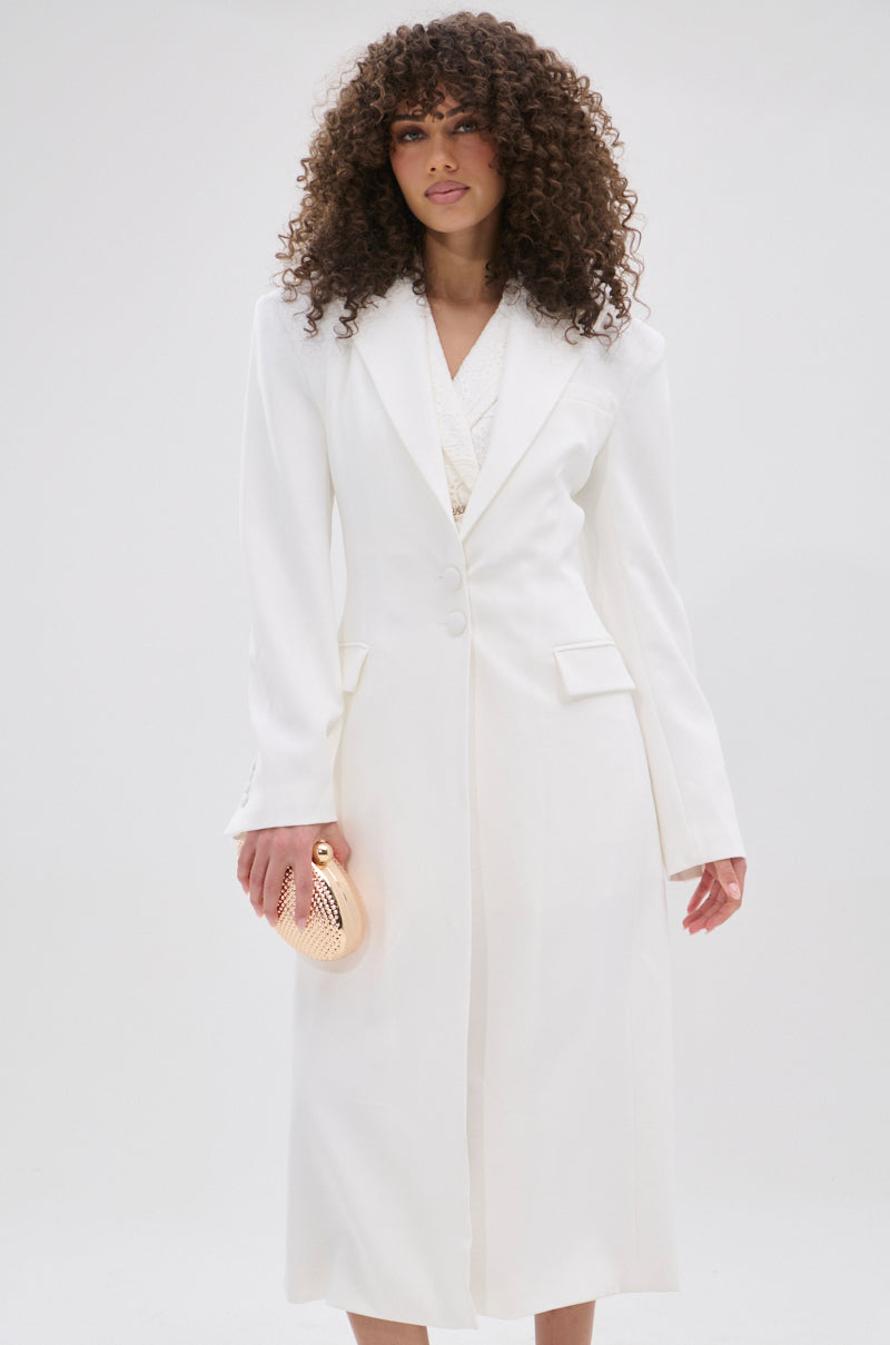 A woman with curly hair wears the PERFECT FIT SLEEK TRENCH in white, styled as a blazer dress. She holds a small gold clutch and stands against a plain light background, looking ahead with confidence.