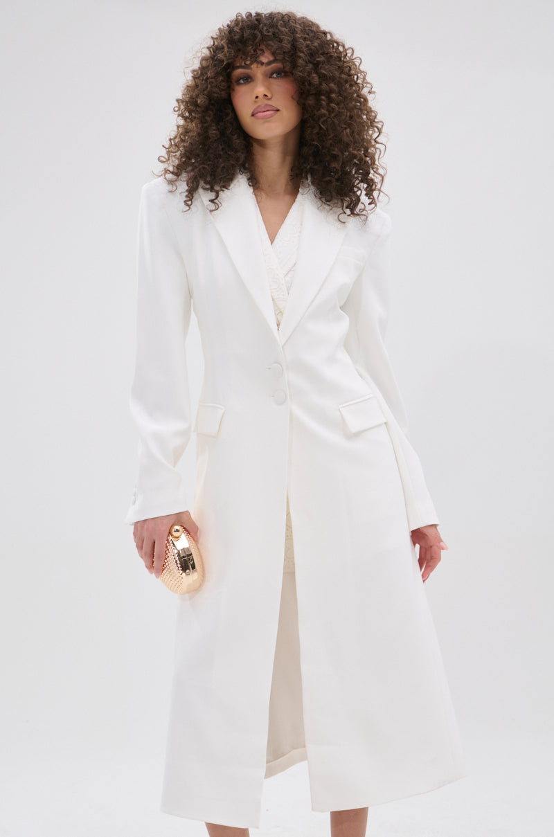 A woman with curly hair wears the PERFECT FIT SLEEK TRENCH—a long, white tailored coat with lapels and buttons—while holding a gold clutch against a plain, light background.