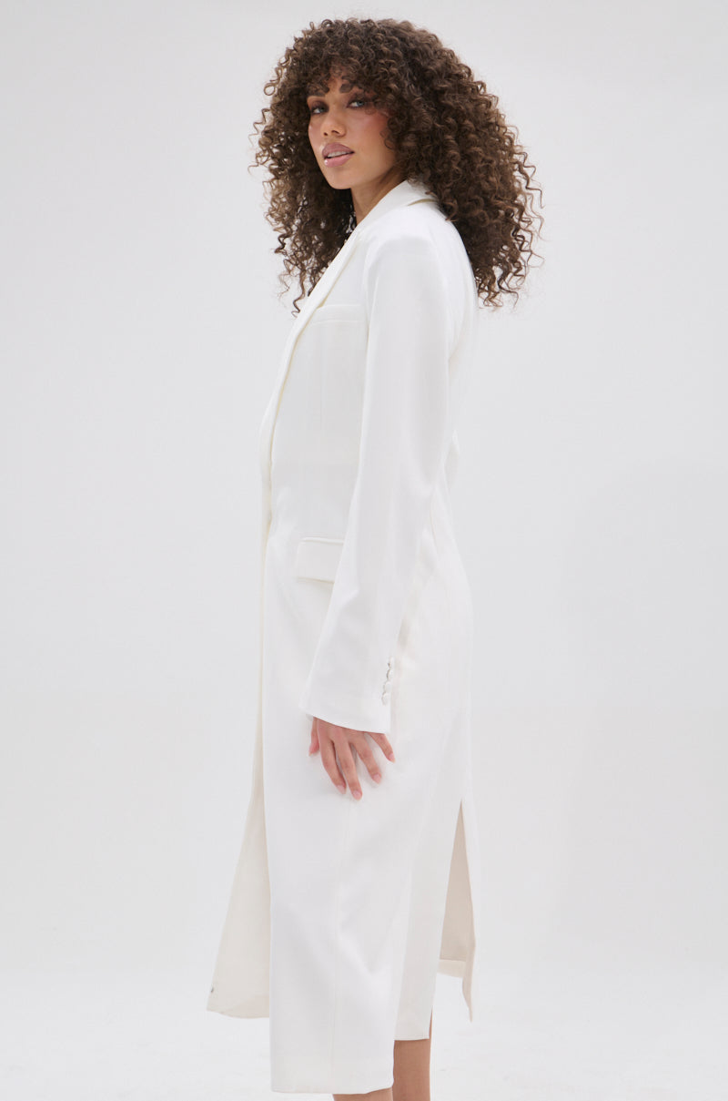 A woman with curly hair models the PERFECT FIT SLEEK TRENCH—a long white coat—standing sideways against a plain light background, looking at the camera with a slight smile and radiating minimalist sophistication.