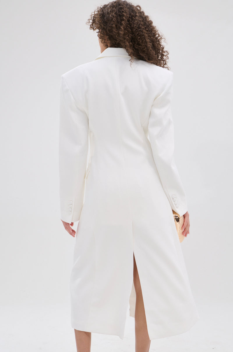 A person with curly hair wears the PERFECT FIT SLEEK TRENCH, a long, white coat with structured shoulders and a center slit, showcasing minimalist sophistication against a plain, light-colored background.