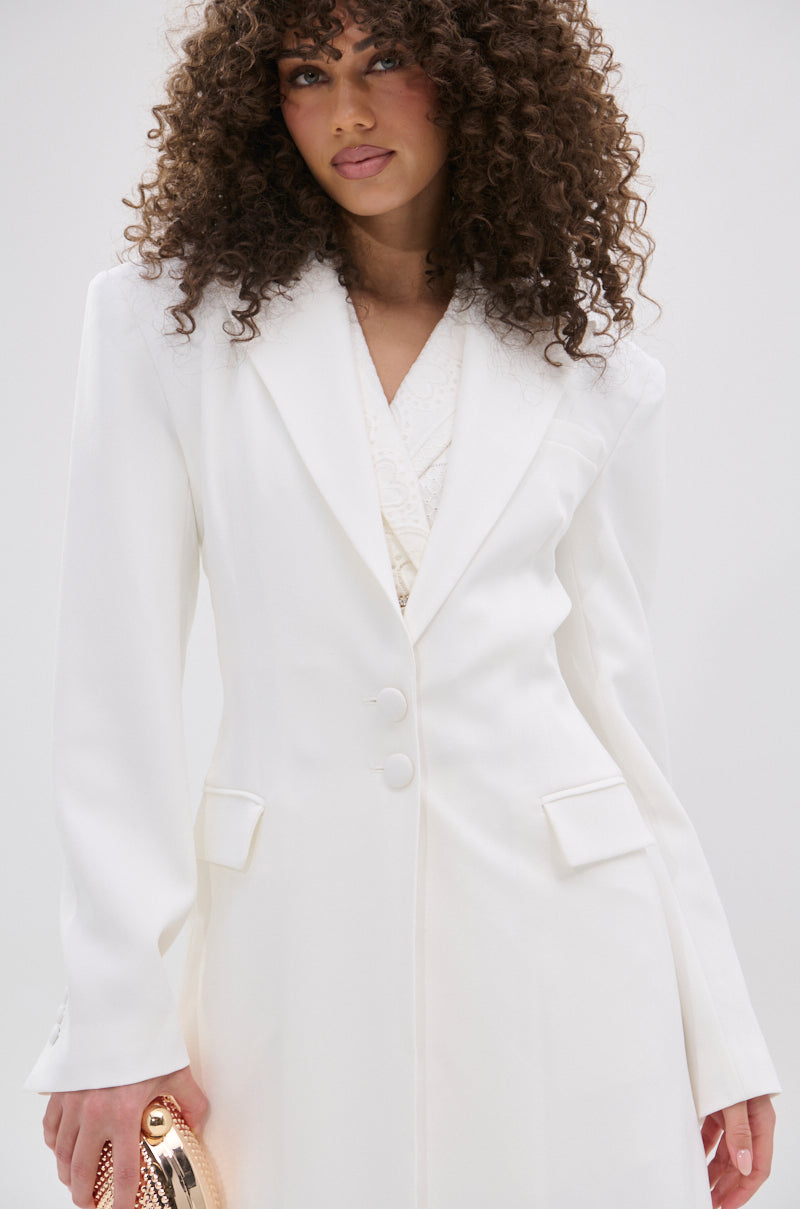 A woman with curly hair wears the PERFECT FIT SLEEK TRENCH over a light top, holding a metallic clutch bag at her side as she stands against a plain light background.