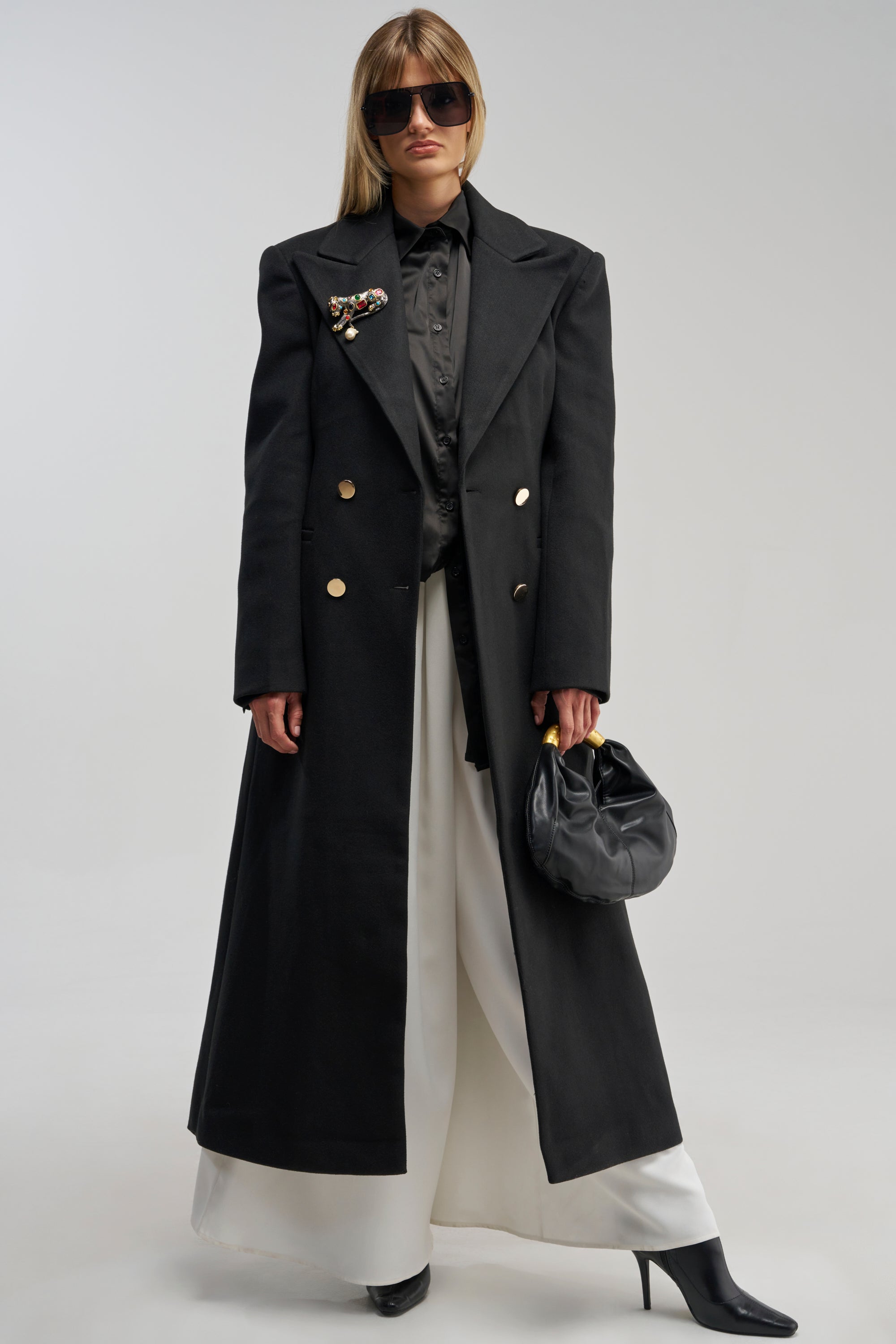 A woman wearing the SCARLETT LUXE TRENCH COAT IN BLACK, styled with a black shirt, white wide-leg pants, black boots, dark sunglasses, and a black handbag stands against a plain background.