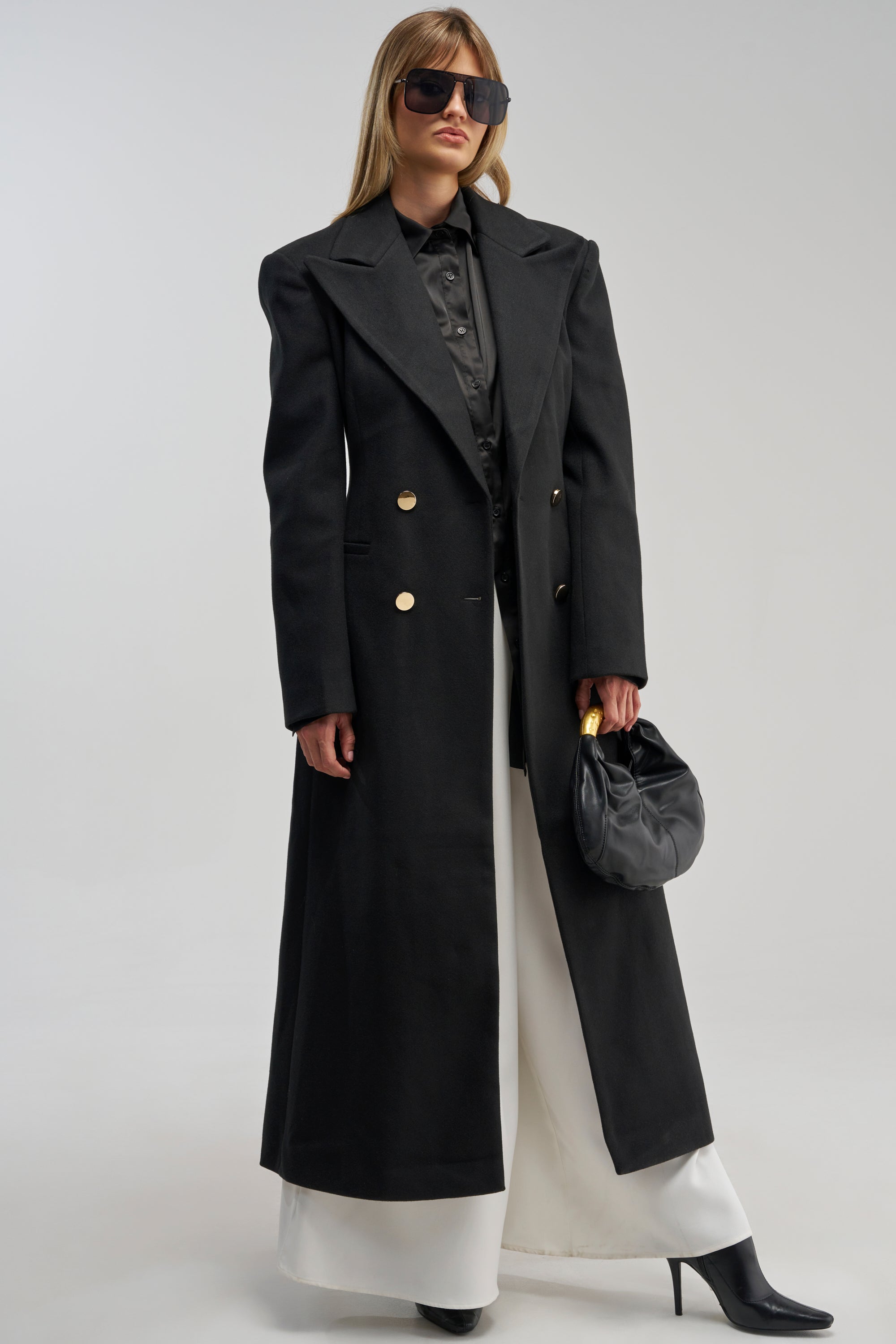 A woman wearing the SCARLETT LUXE TRENCH COAT IN BLACK, styled with a black shirt, white wide-leg pants, black heels, sunglasses, and carrying a small black purse stands against a plain light background.