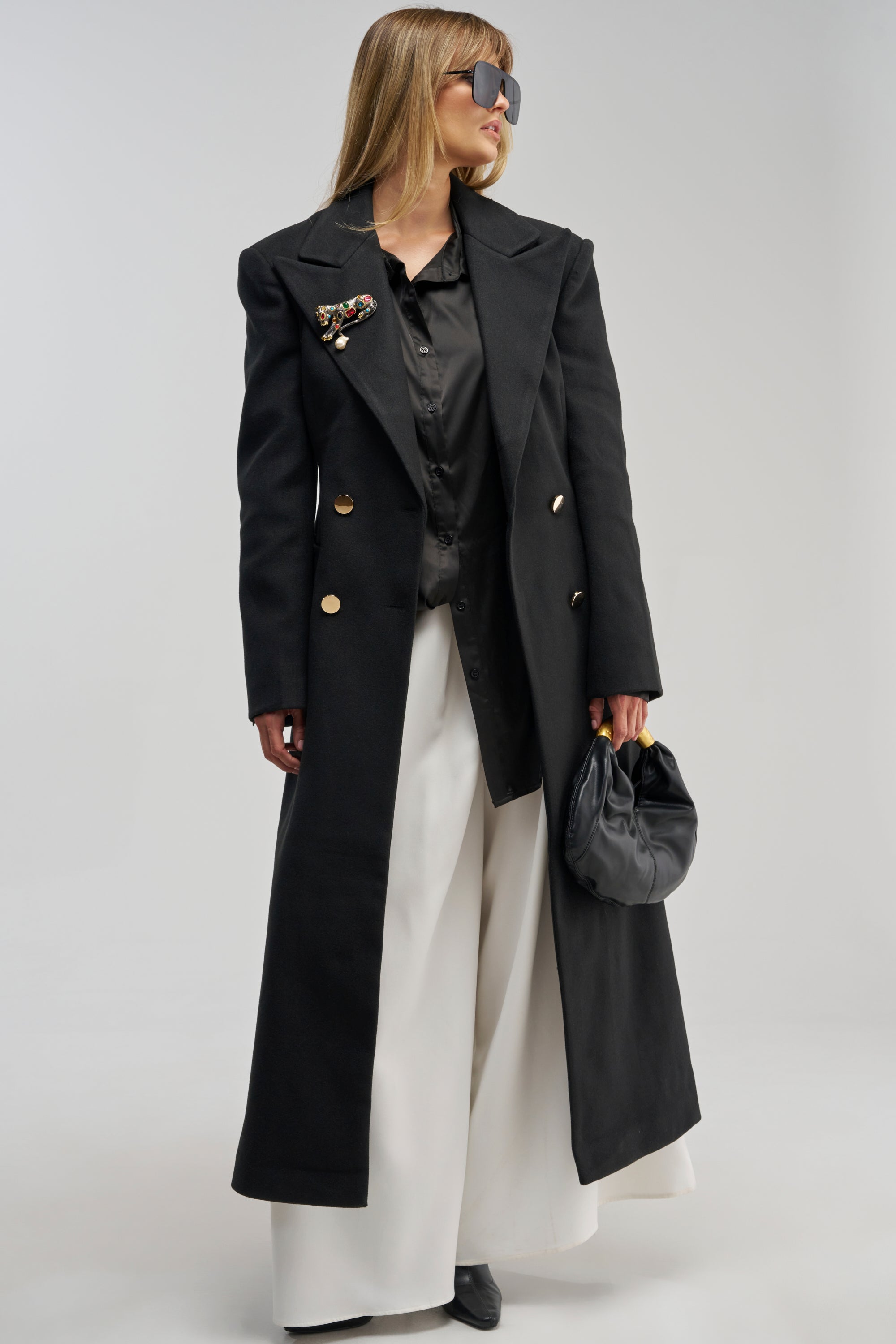 Wearing the SCARLETT LUXE TRENCH COAT IN BLACK with gold buttons, a black shirt, wide white pants, sunglasses, and holding a small black bag, a woman stands against a plain background.