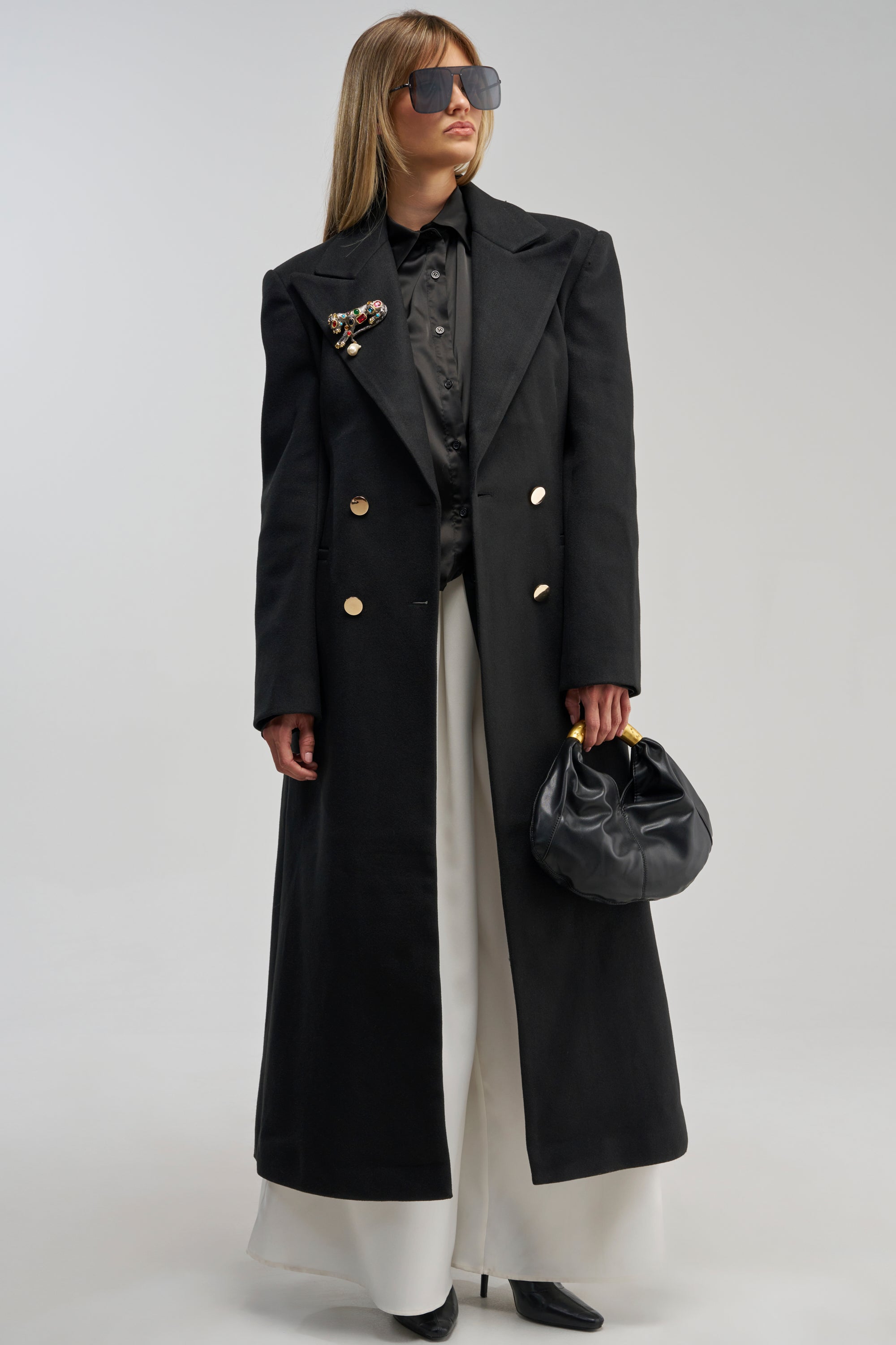 A person wearing the SCARLETT LUXE TRENCH COAT IN BLACK, styled with gold buttons, white wide-leg pants, black boots, sunglasses, and holding a small black handbag stands against a plain light background.