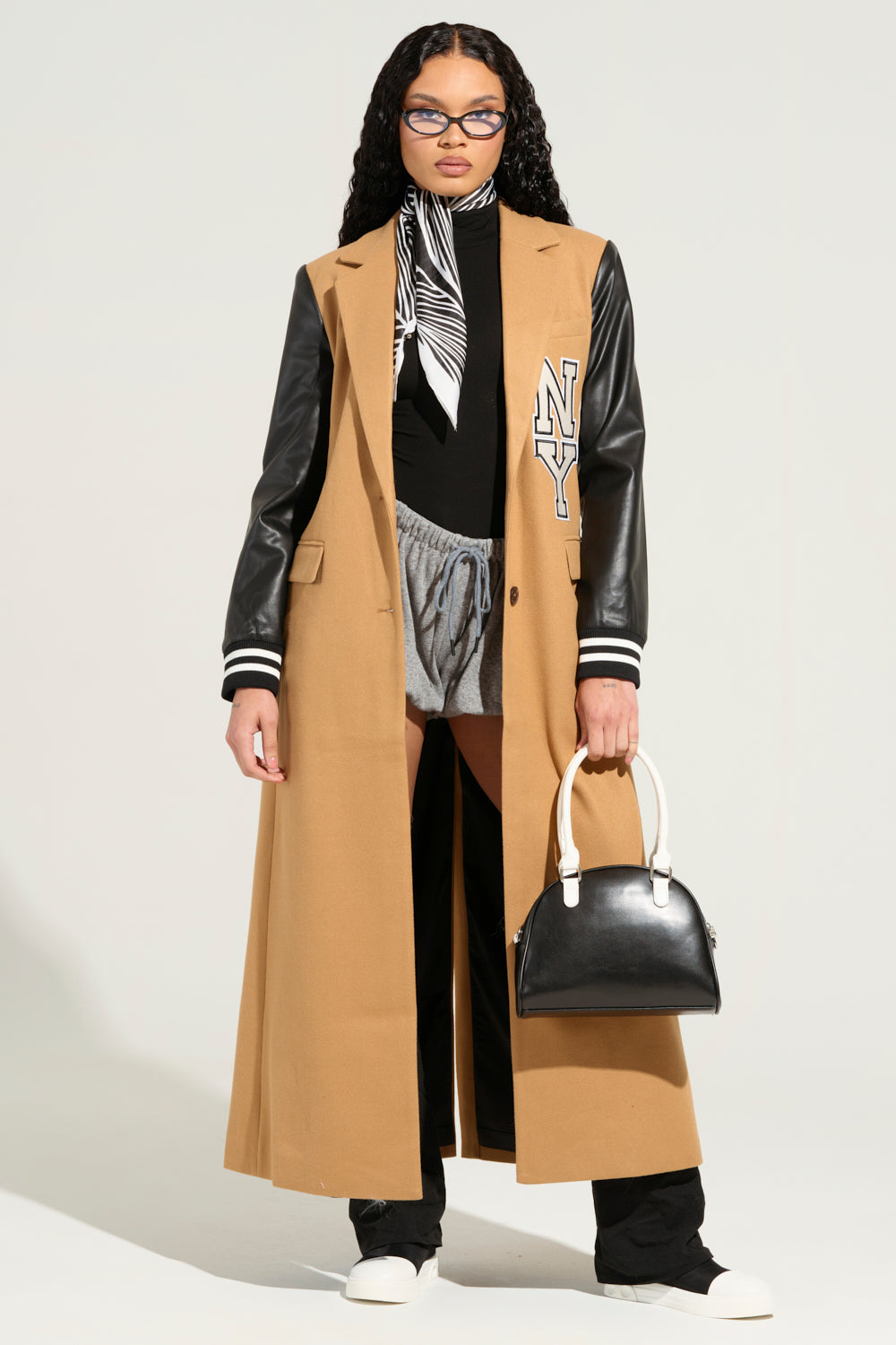 A person is dressed in the BRENTWOOD 2.0 VARSITY TRENCH with black faux leather sleeves displaying an "NY" logo, paired with a black top, gray pants, and white shoes. They are carrying a black and white handbag, accessorized with a zebra-patterned scarf, and have long curly hair and glasses.