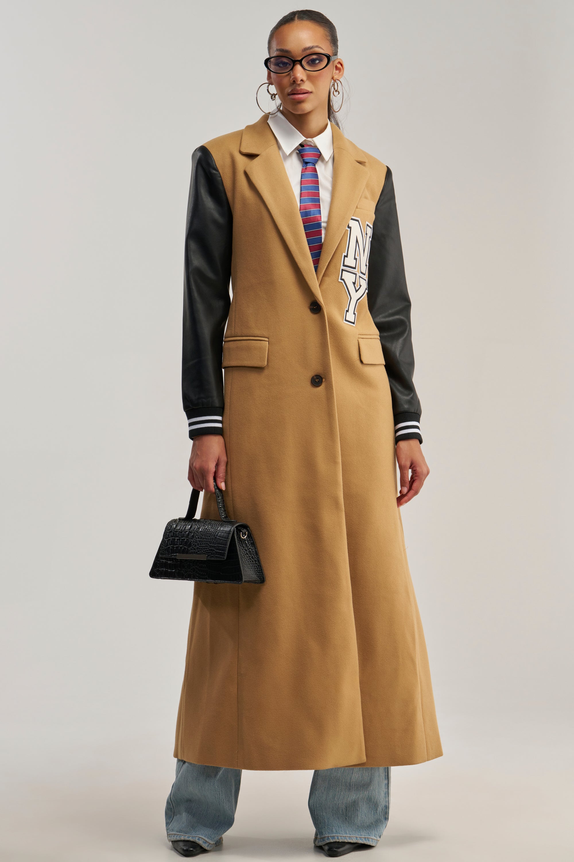 Wearing the BRENTWOOD 2.0 VARSITY TRENCH—a tan trench coat with faux leather sleeves and an “NY” patch—this person channels a streetwear vibe, paired with blue jeans, a striped tie, glasses, hoop earrings, and a small black handbag.