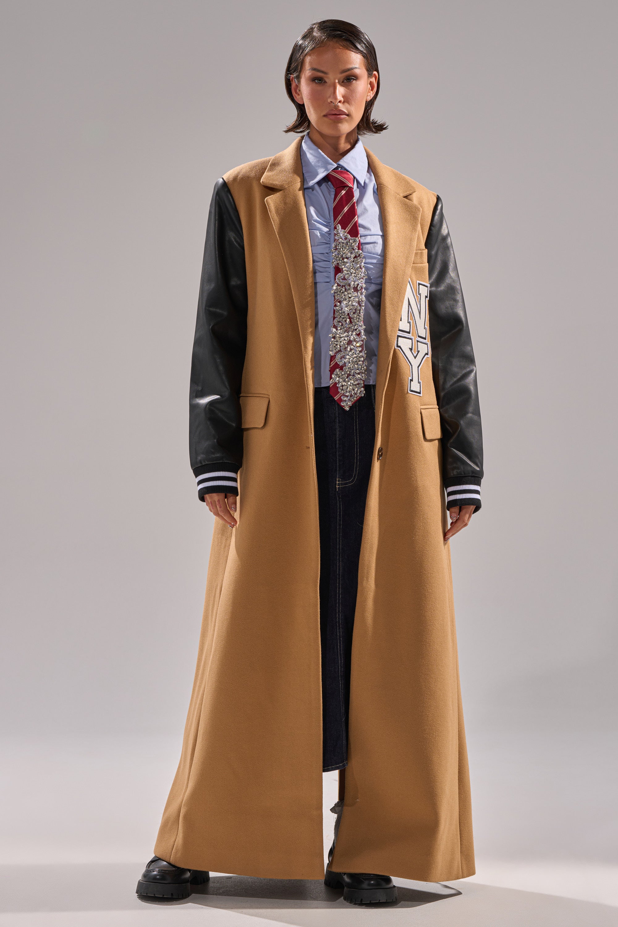 A person faces forward wearing the BRENTWOOD 2.0 VARSITY TRENCH with black faux leather sleeves, an "NY" patch on the left, over a blue shirt, silver tie, dark pants, and black shoes. The coat features a longline varsity silhouette.