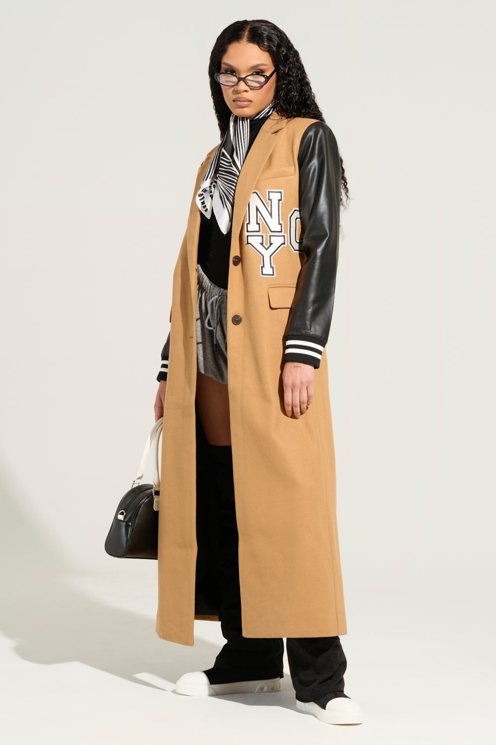 A stylish person showcases a chic BRENTWOOD 2.0 VARSITY TRENCH with "NY" on the chest and faux leather sleeves. They're wearing white sneakers and sunglasses, carrying a black bag. A patterned scarf adorns their striped shirt, paired with gray shorts over black leggings.