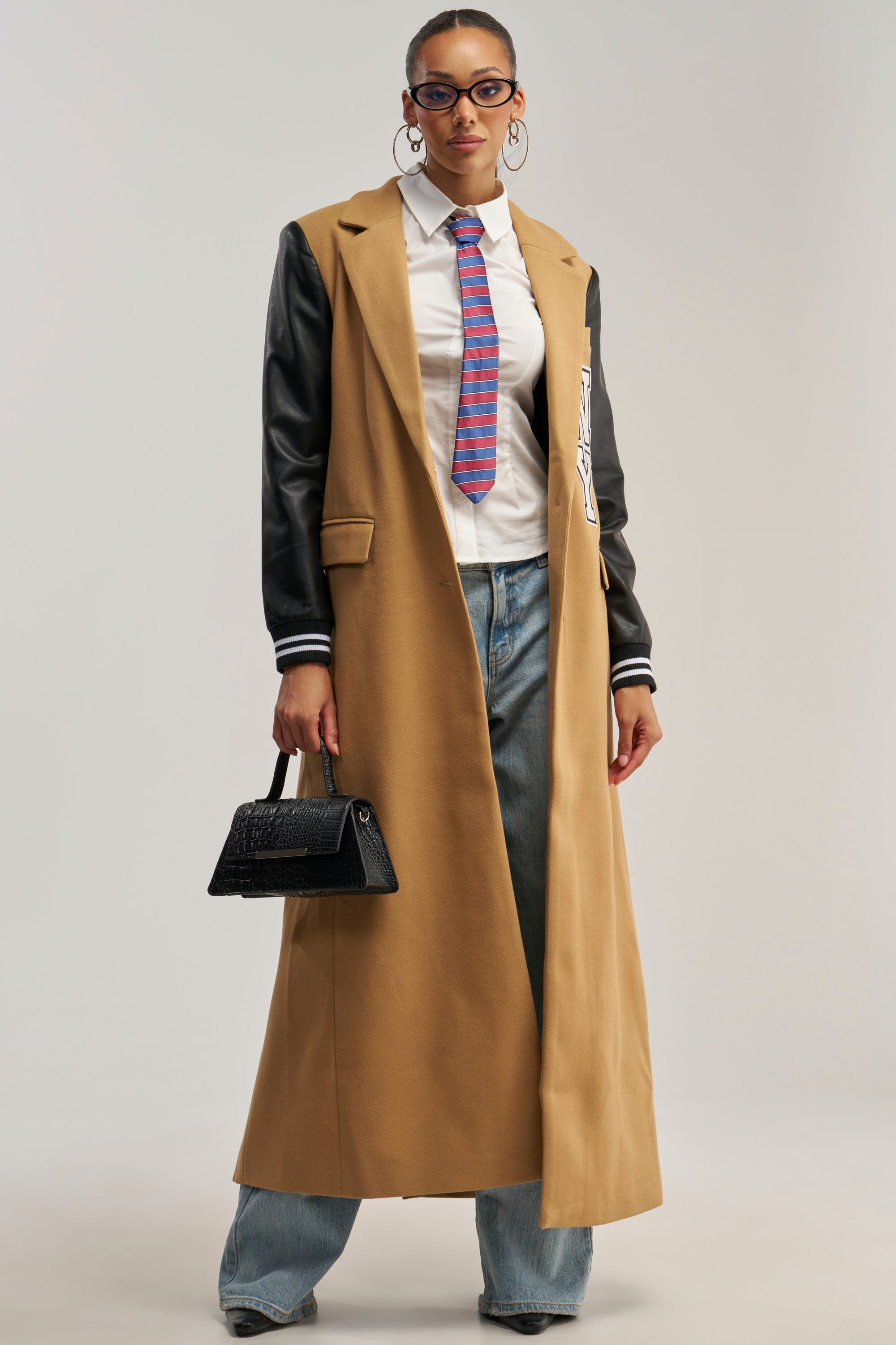 A woman stands confidently against a plain background, embodying chic streetwear in the BRENTWOOD 2.0 VARSITY TRENCH—a long tan coat with faux leather sleeves—styled with sunglasses, hoop earrings, a white shirt, colorful tie, blue jeans, and a black handbag.