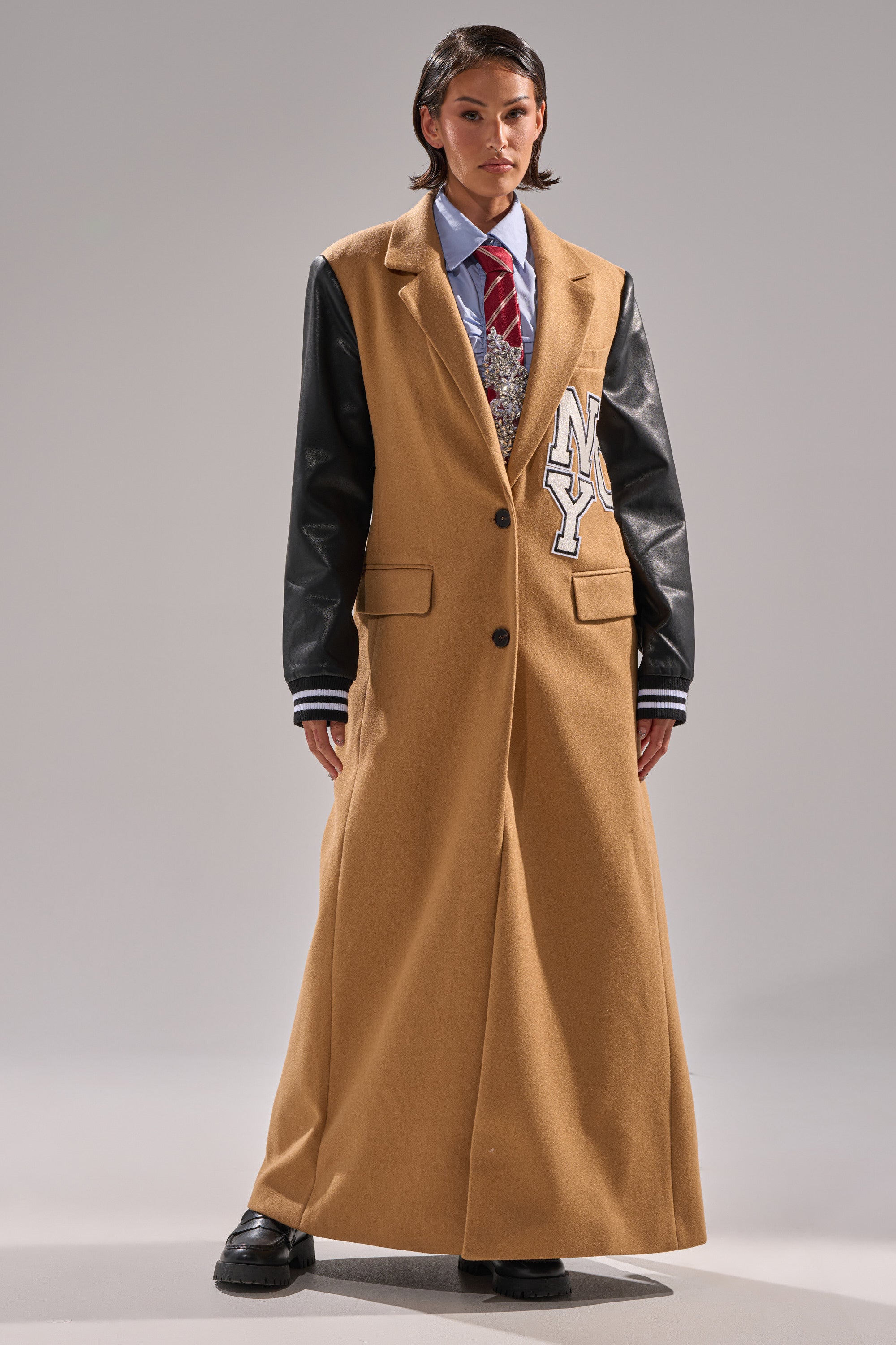 A person stands against a plain background wearing the BRENTWOOD 2.0 VARSITY TRENCH, a long tan coat with faux leather sleeves and an NY patch, layered over a shirt, tie, patterned sweater, and paired with black shoes.
