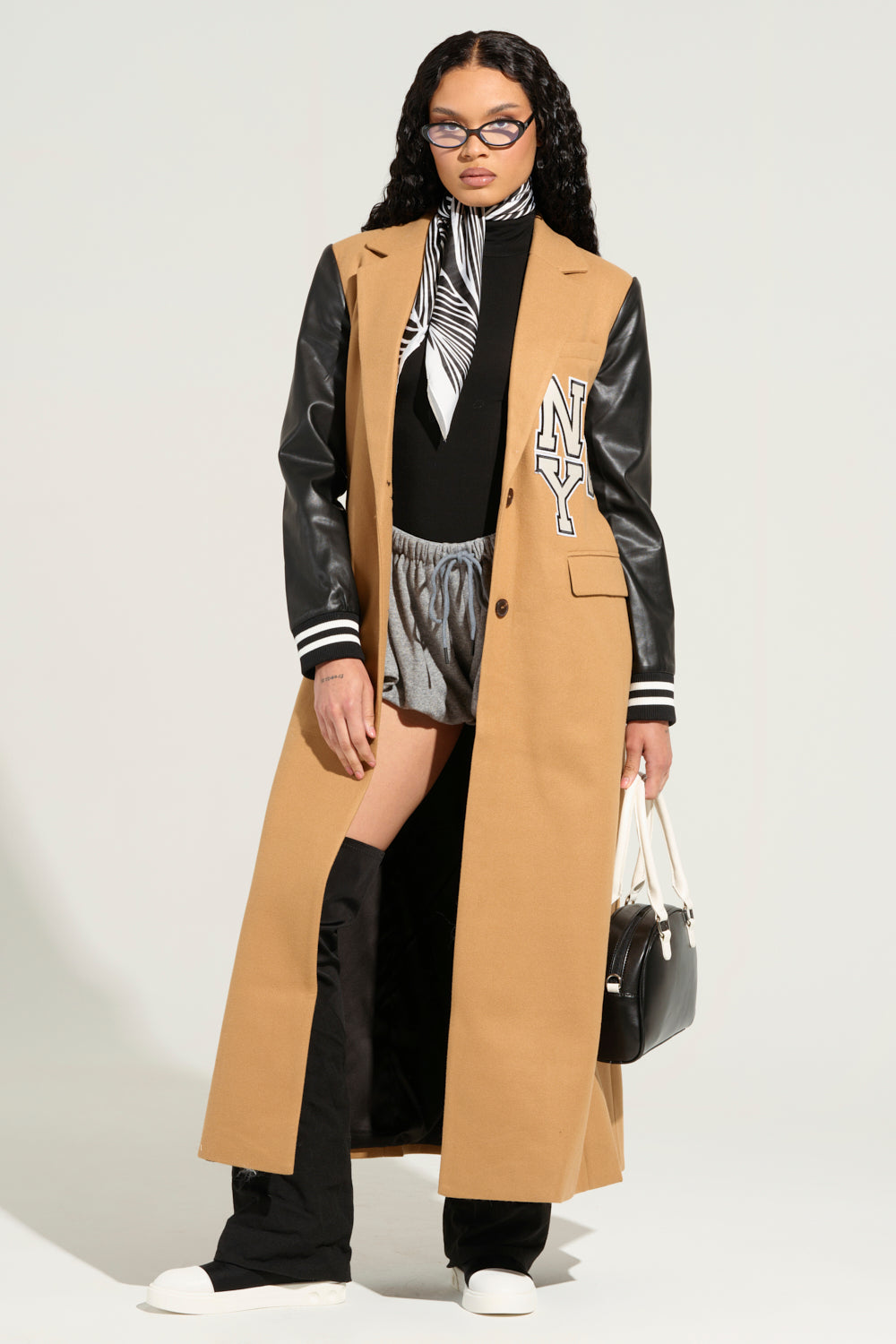 A person wearing the BRENTWOOD 2.0 VARSITY TRENCH with faux leather sleeves and a black turtleneck, paired with gray shorts and knee-high black boots. They accessorize with glasses, a stylish black and white handbag, and a matching scarf around their neck for an effortlessly chic look.