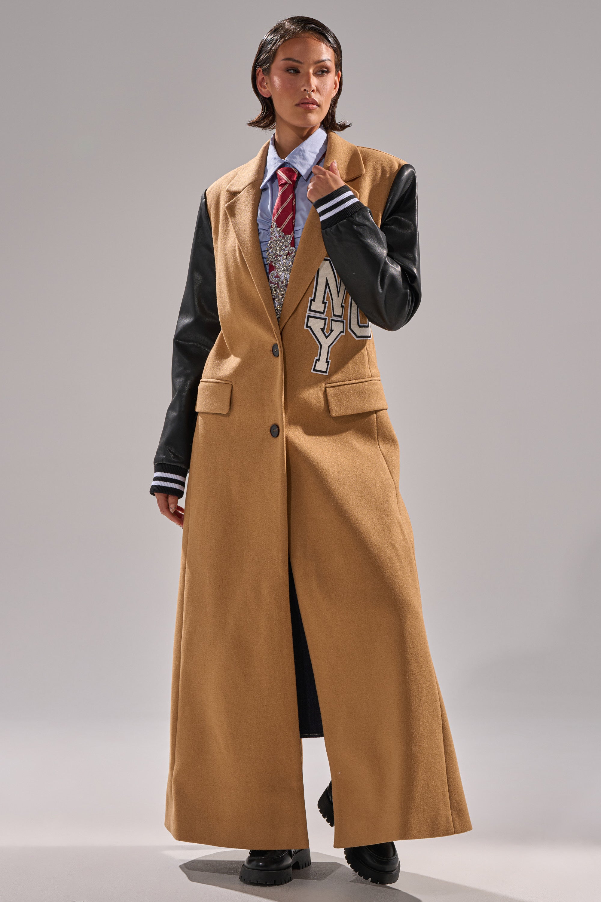 A person in the BRENTWOOD 2.0 VARSITY TRENCH, a tan coat with faux leather sleeves and a large white "NY" patch, paired with a blue shirt, red striped tie, and black shoes.
