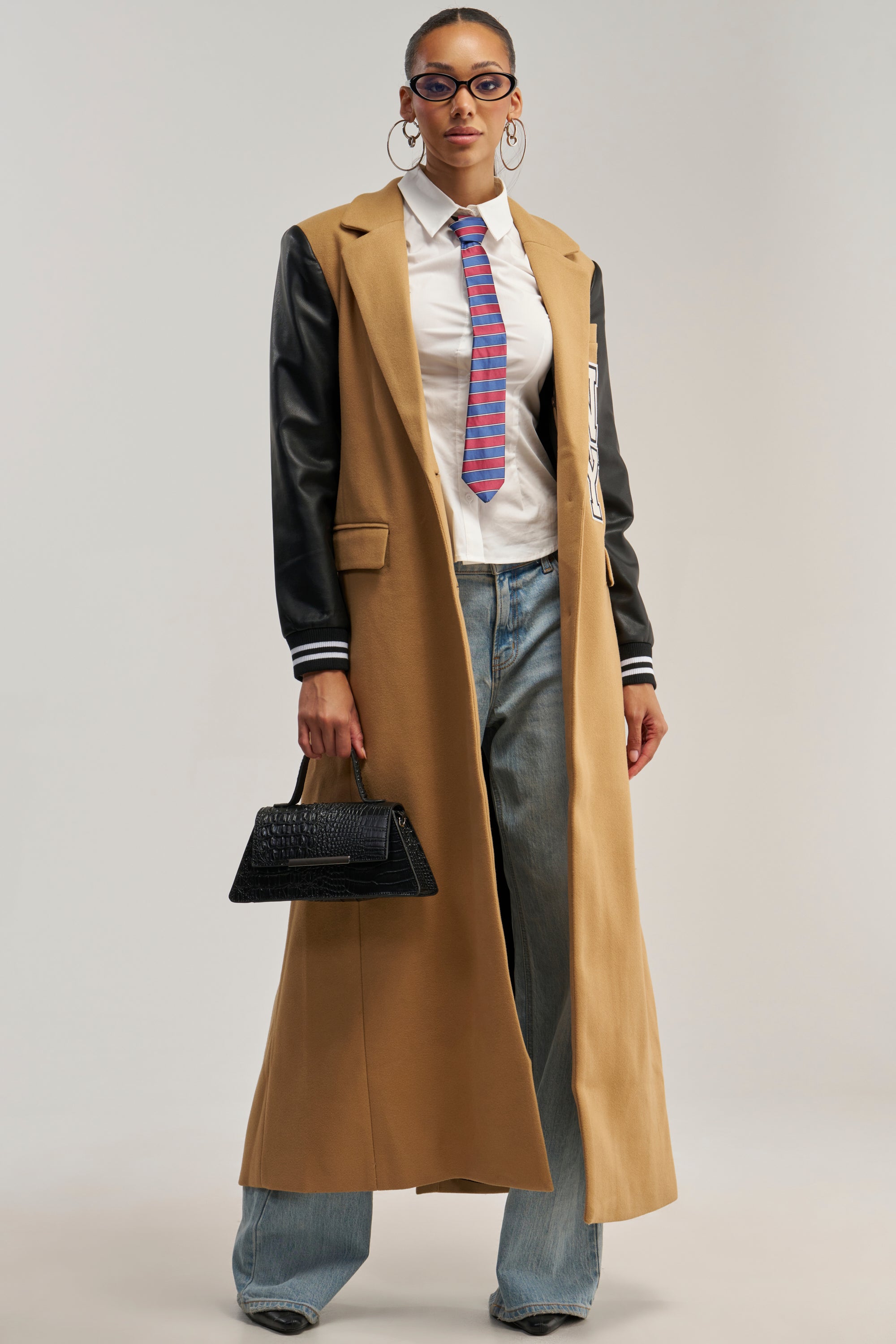Wearing the BRENTWOOD 2.0 VARSITY TRENCH, a woman exemplifies streetwear style with its tan design featuring faux leather sleeves, paired with a crisp white shirt, striped tie, loose jeans, sunglasses, hoop earrings, and a black handbag.