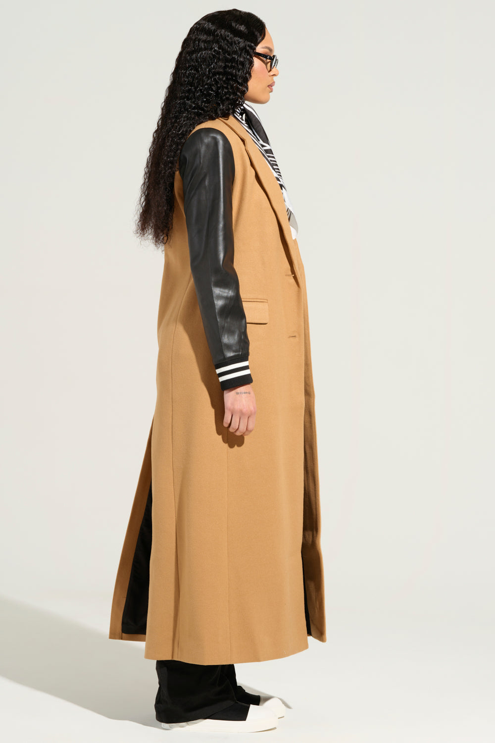 A person with long, curly hair stands in profile wearing glasses and the BRENTWOOD 2.0 VARSITY TRENCH, which features black faux leather sleeves, styled over a black and white striped shirt. They complete the look with black pants and white shoes against a plain light-colored background.