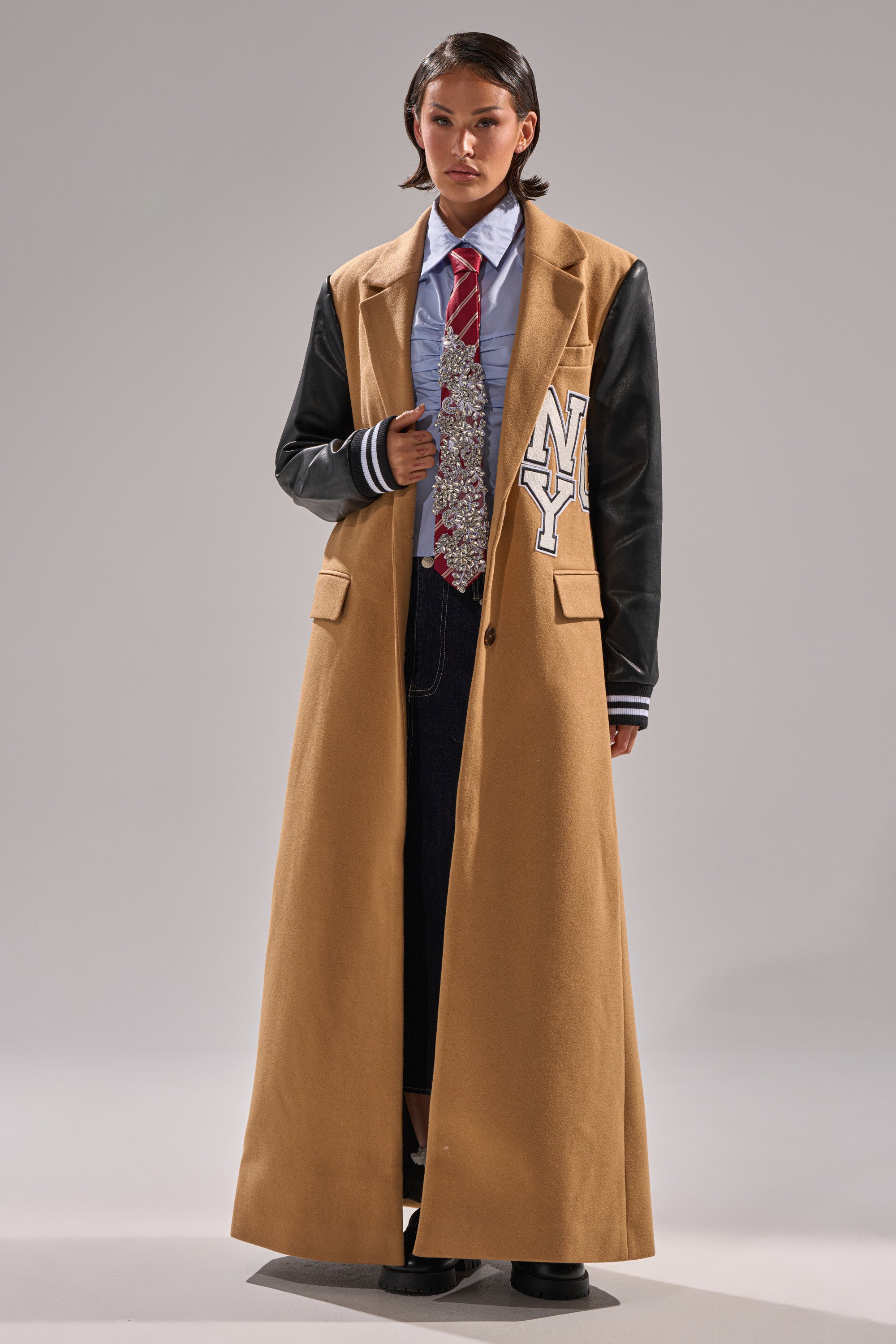 A person wears the BRENTWOOD 2.0 VARSITY TRENCH featuring faux leather sleeves and large letter patches, paired with a blue button-up, red striped tie, dark pants, and black shoes. The coat is floor-length. Background: plain gray.