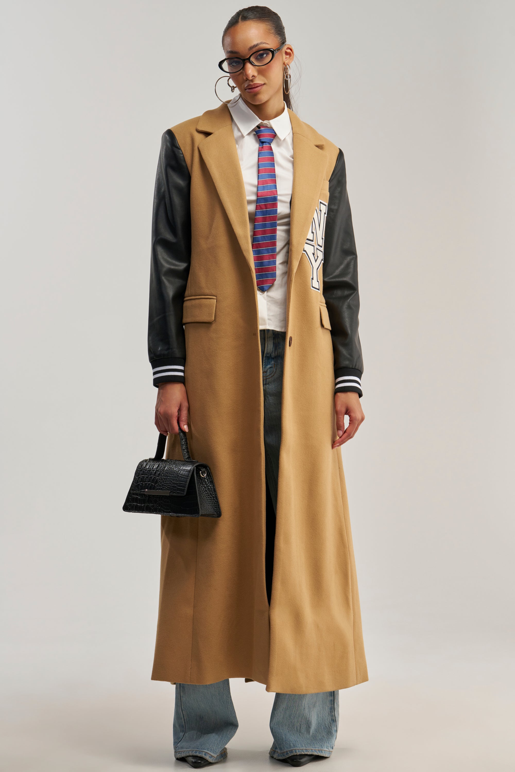 A person stands against a neutral background wearing the BRENTWOOD 2.0 VARSITY TRENCH—a long tan trench coat with faux leather sleeves—paired with light blue jeans, a white shirt, a colorful tie, glasses, hoop earrings, and holding a small black handbag. The look blends classic style with modern streetwear influences.