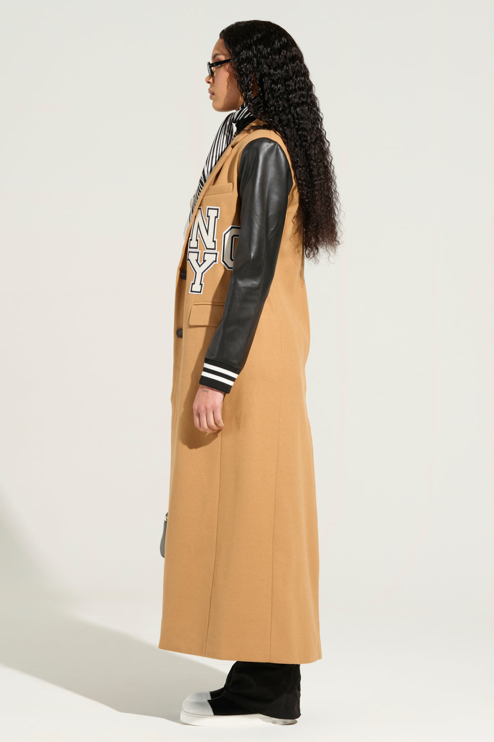 The individual with long curly hair and glasses is wearing the BRENTWOOD 2.0 VARSITY TRENCH, a tan coat highlighted with white letters and complemented by faux leather sleeves. They seamlessly pair it with black pants and white shoes, set against a plain white background.