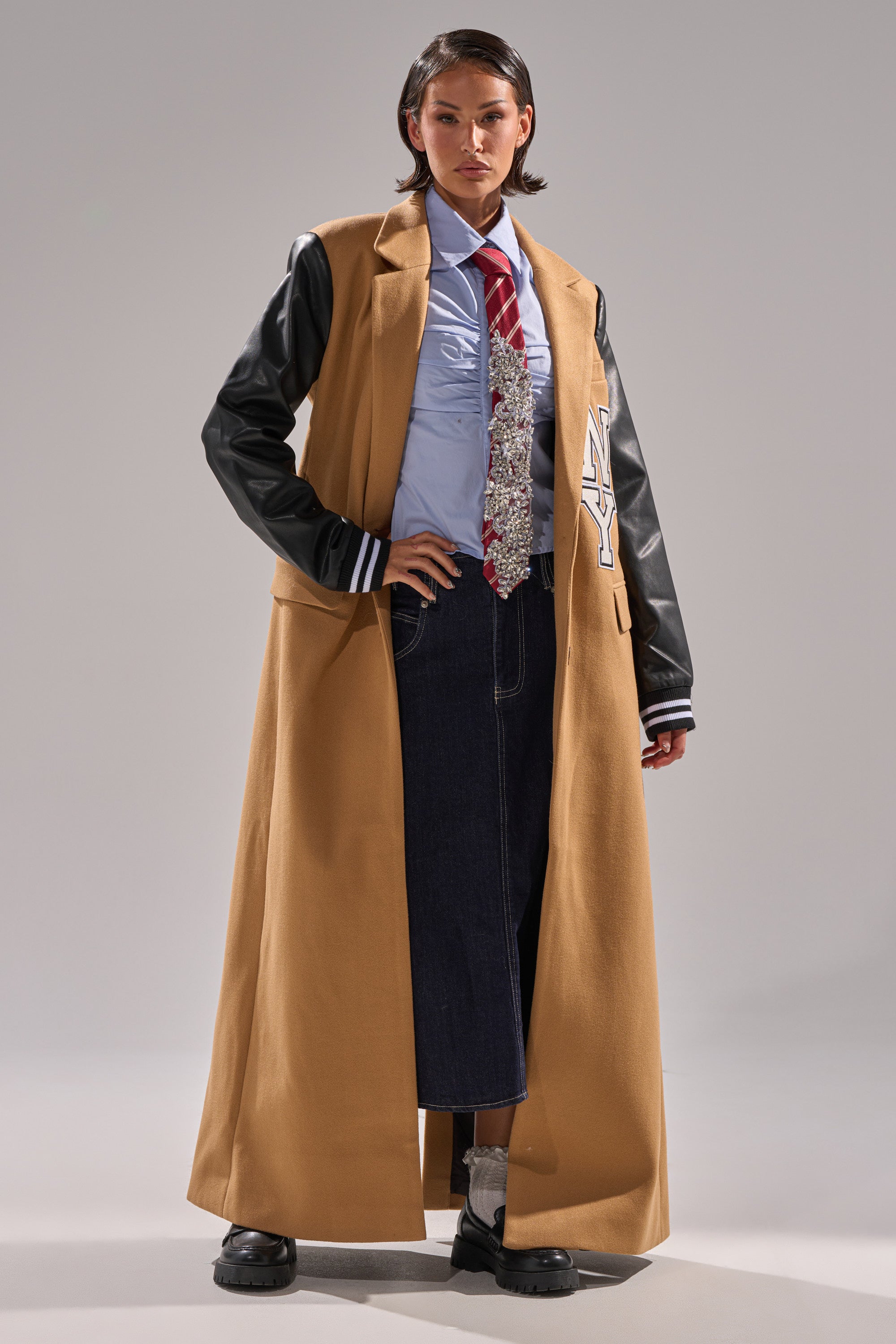 Wearing the BRENTWOOD 2.0 VARSITY TRENCH with faux leather sleeves, a blue shirt, red striped tie, dark wide-leg jeans, and chunky black shoes, this look blends formal and casual styles with confidence.