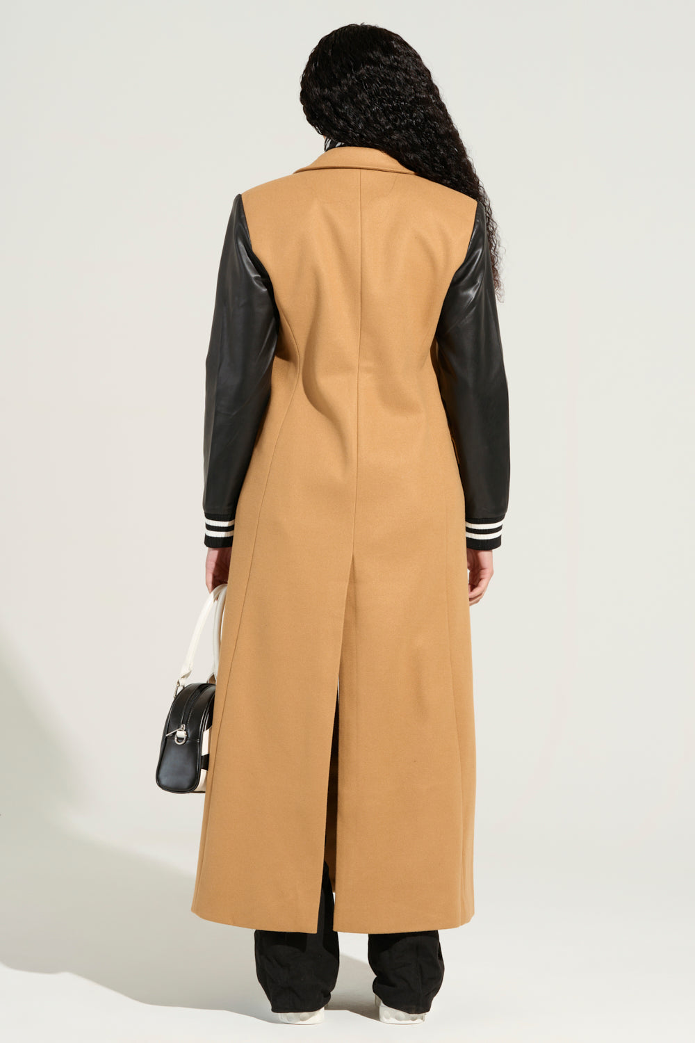 A person with long braided hair stands facing away, dressed in the BRENTWOOD 2.0 VARSITY TRENCH with faux leather sleeves and a slit at the back. They carry a black and white handbag while wearing black pants against a plain background.