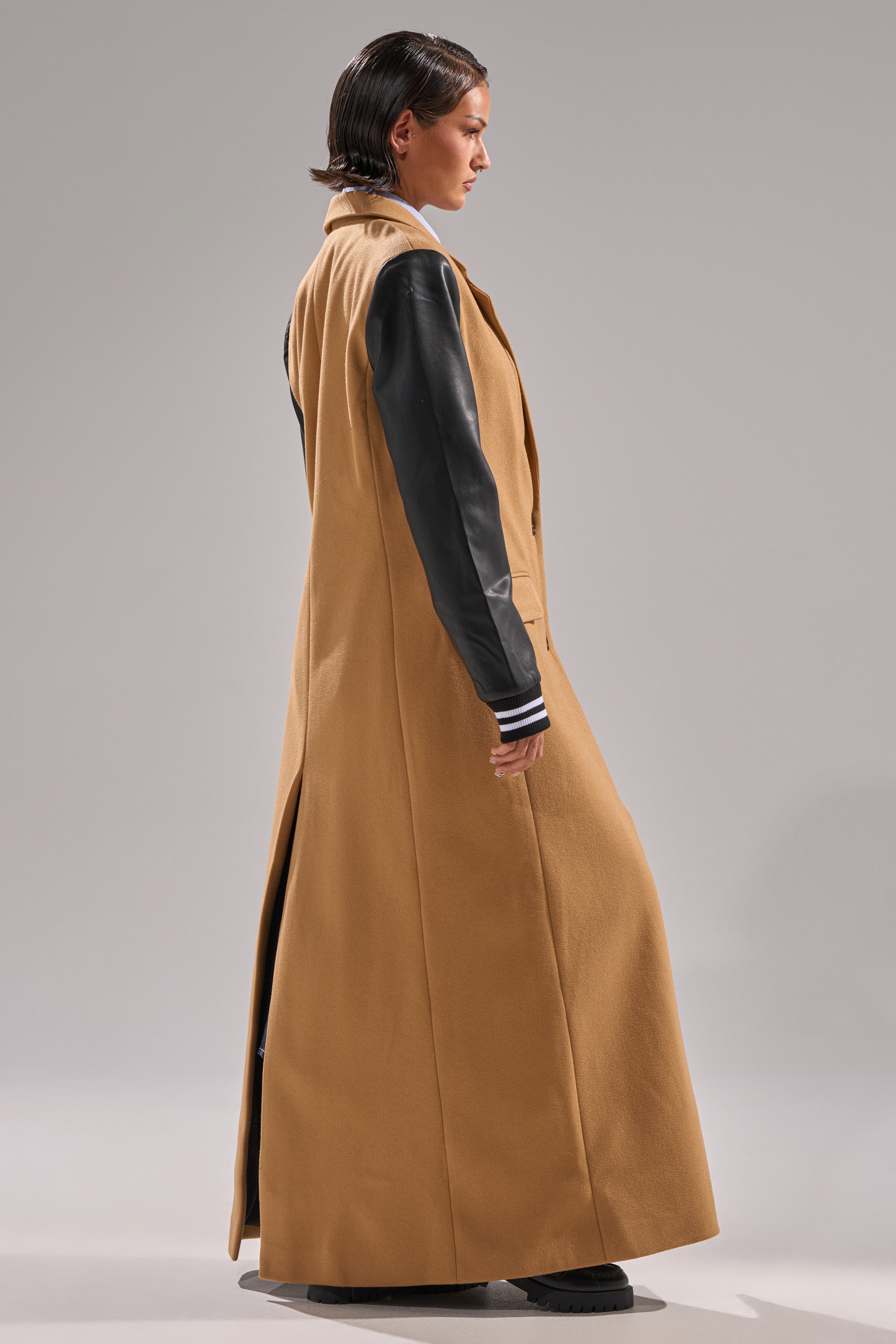 A person stands sideways modeling the BRENTWOOD 2.0 VARSITY TRENCH—a long tan coat with black leather sleeves, a back slit, and black shoes—against a plain light gray background.