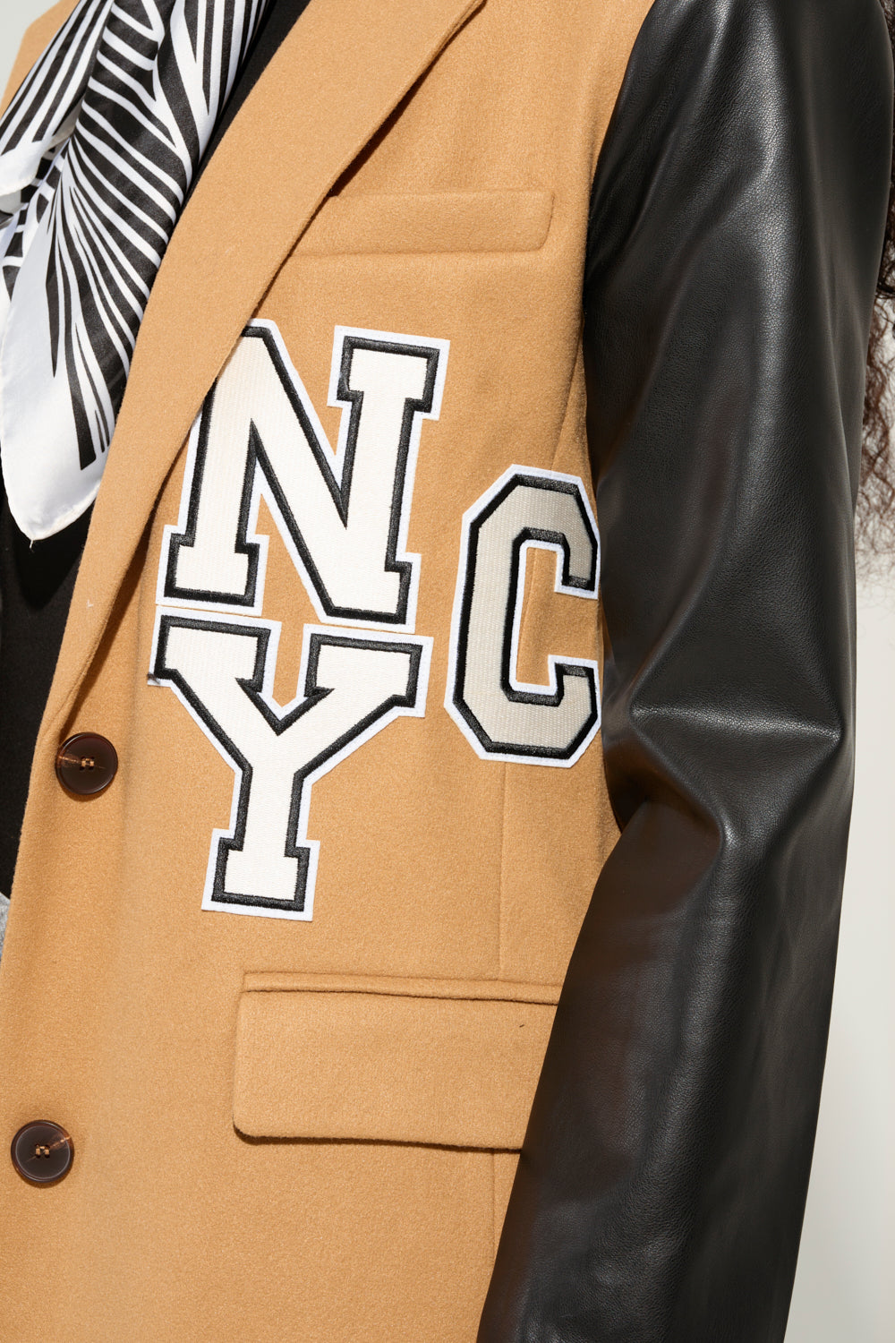 Close-up of the BRENTWOOD 2.0 VARSITY TRENCH in tan, featuring prominent "NYC" letters on the chest and faux leather sleeves. A person partially visible in the background is accessorized with a black and white striped scarf, enhancing this stylish scene.
