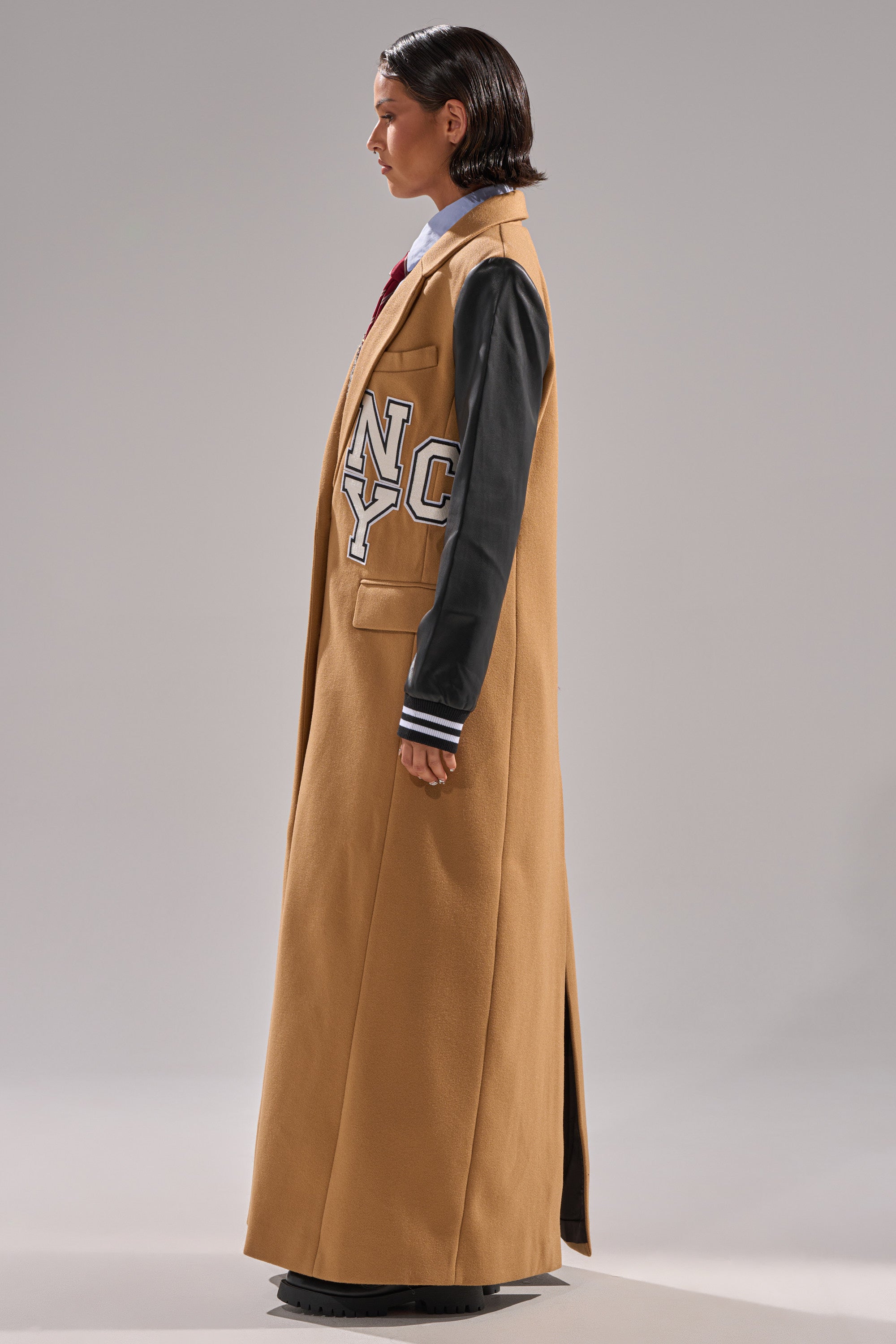 A person stands in profile against a plain grey background, wearing the BRENTWOOD 2.0 VARSITY TRENCH with black faux leather sleeves, varsity-style patches, and a white collared shirt underneath.