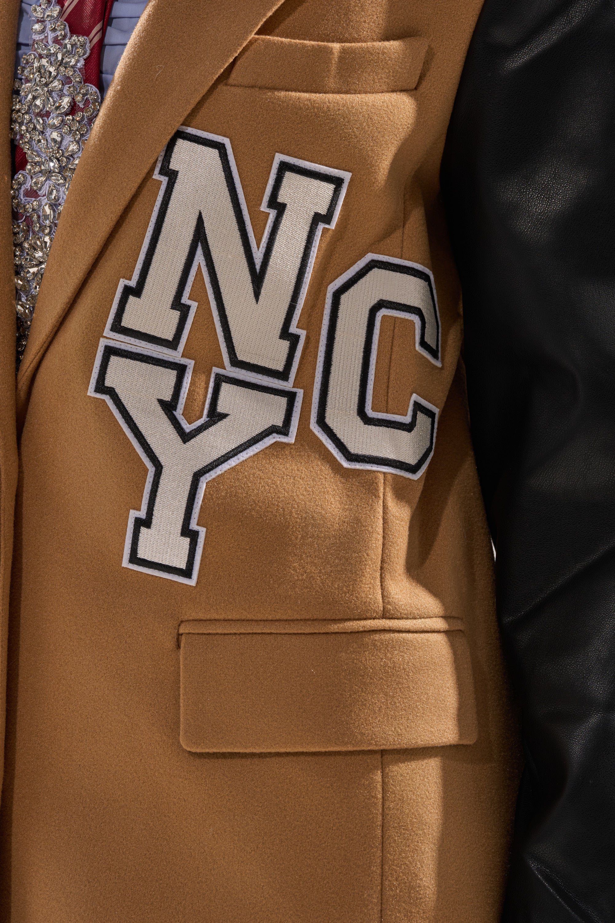 Close-up of the BRENTWOOD 2.0 VARSITY TRENCH—a brown coat with black faux leather sleeves and a black and white "NYC" patch on the chest, styled over a patterned shirt and tie. Part of the right sleeve is shown.