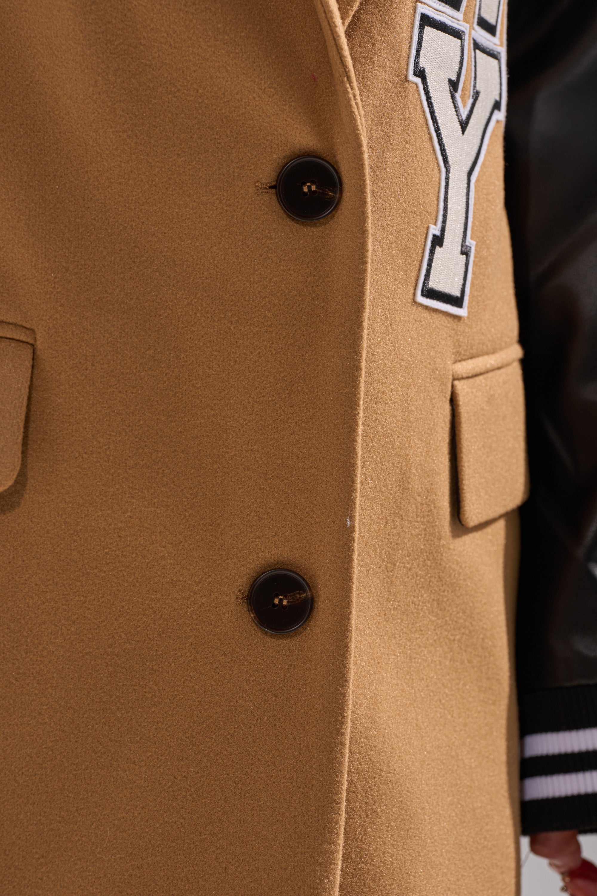 Close-up of the BRENTWOOD 2.0 VARSITY TRENCH: tan body, black buttons, large white "Y" patch, faux leather sleeves, striped cuff, and front pocket visible on the right.