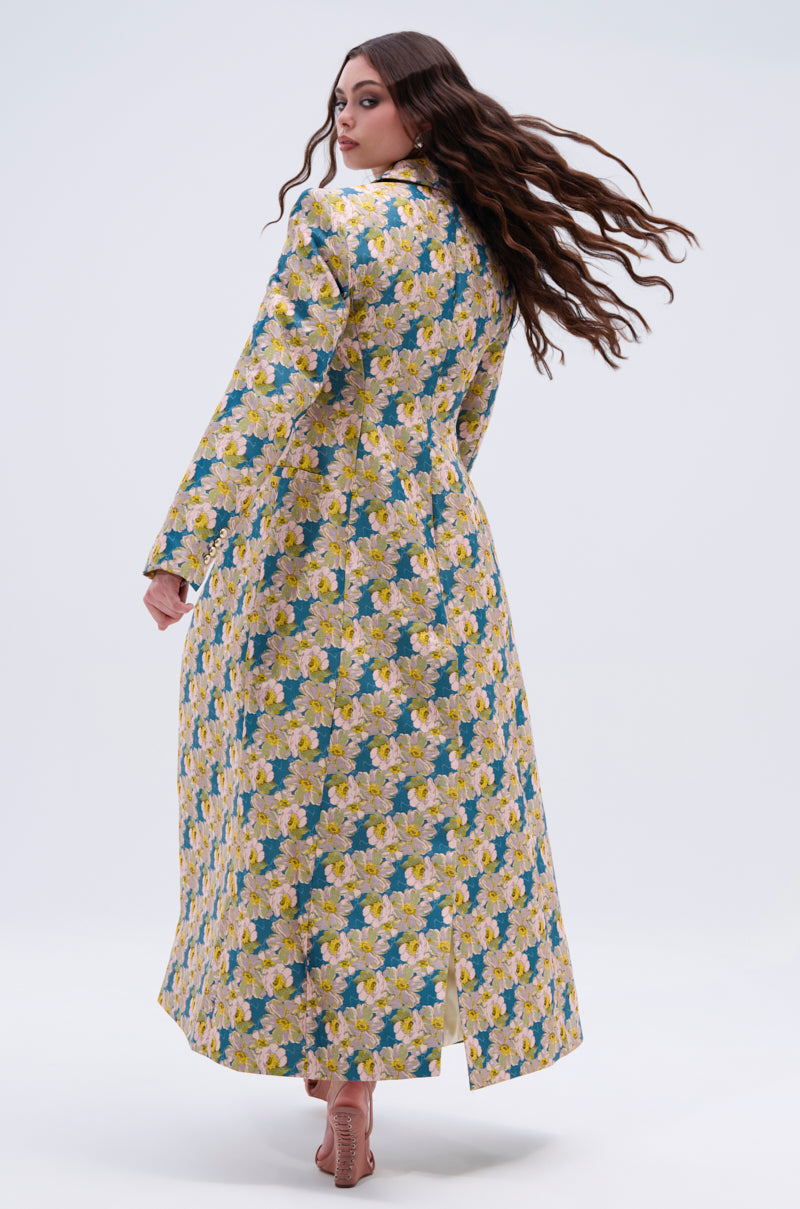 A woman with long, wavy brown hair models the SCARLETT LUXE TRENCH IN BROCADE, a floral-patterned coat in yellow and blue. Barefoot, she looks over her shoulder against a plain light background.